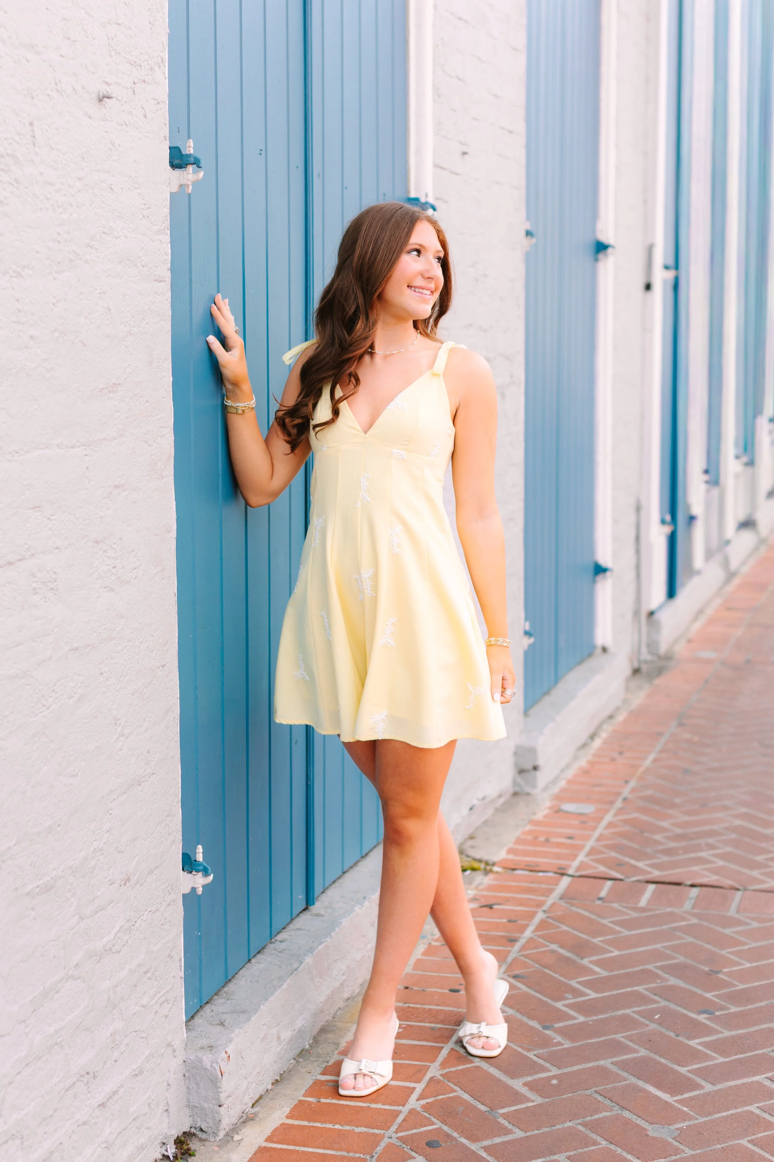 A young woman in a yellow dress and white heels standing by a wall with blue doors, smiling and looking to the side.