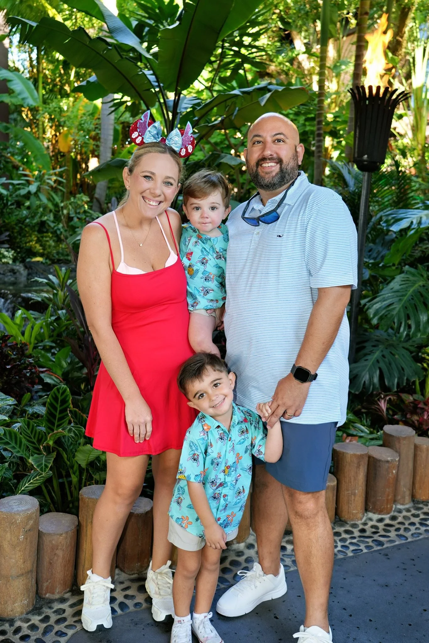 A family of five smiling and posing outdoors in a lush tropical garden, with green foliage and decorative torch lights in the background. The woman is dressed in a red dress with white sneakers, wearing a headband with red, white, and blue decorations. The man is wearing a light blue striped polo shirt and navy shorts, with sunglasses hanging from his neck. The two young boys are in matching tropical-themed shirts; one is holding hands with the man, and the other is standing in front of the woman, all smiling.