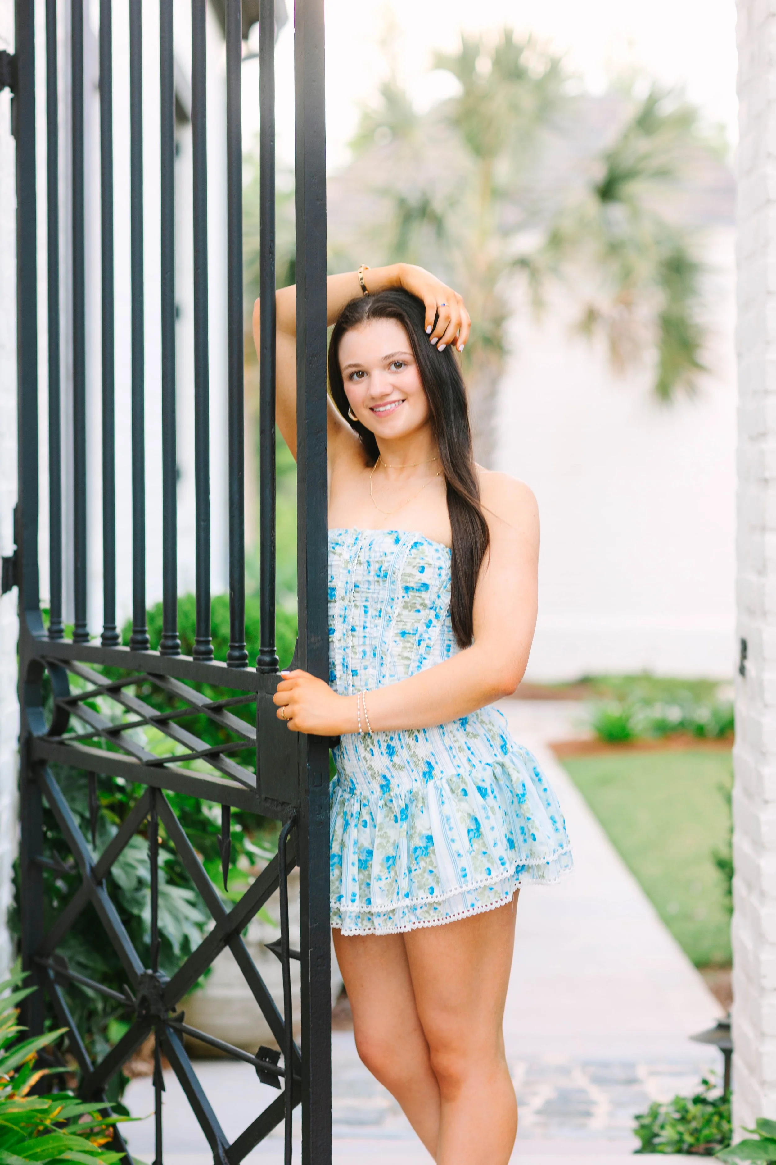 Young woman in a blue and white patterned summer dress smiling and posing behind a black metal gate, with a garden and palm trees in the background.