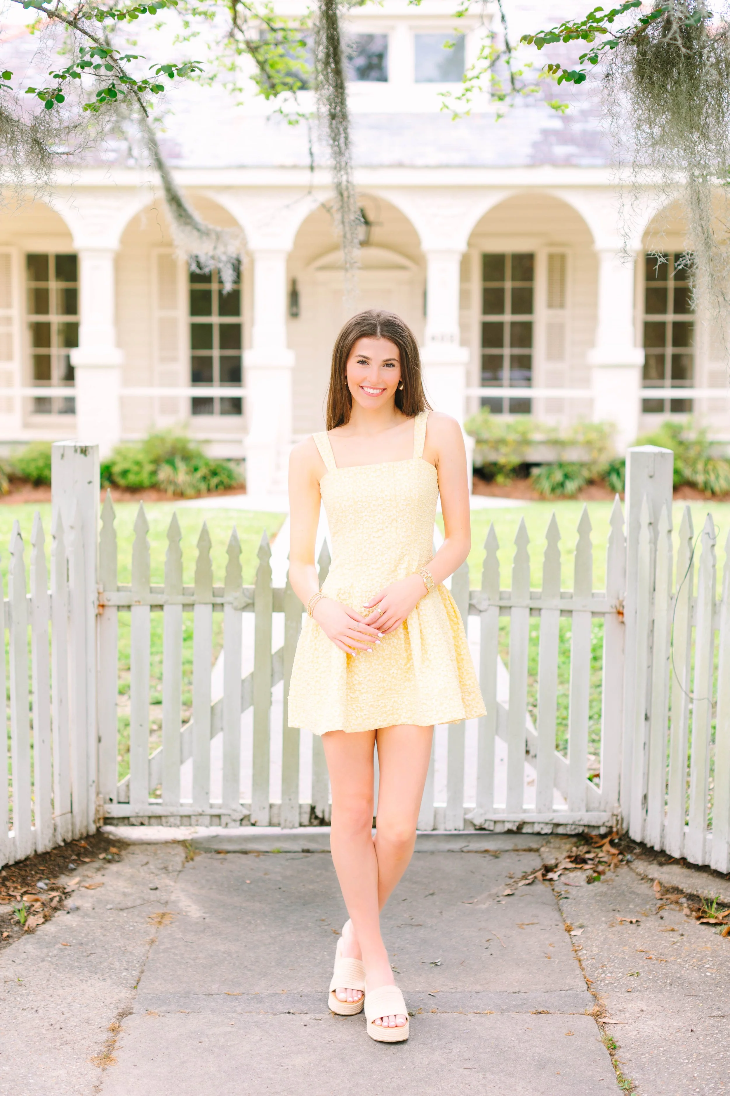 A young woman stands on a sidewalk in front of a white picket fence, smiling at the camera, wearing a yellow sundress and platform sandals, with a house and greenery in the background.