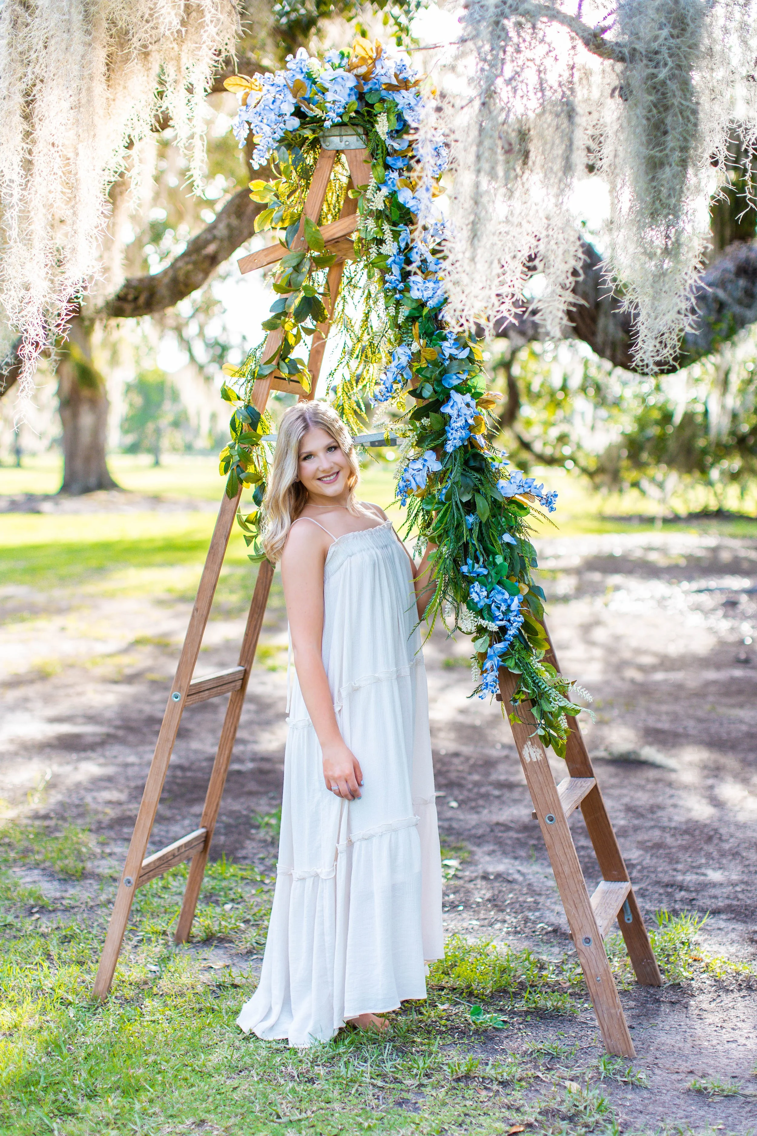 A young woman with wavy blonde hair is smiling at the camera while standing in front of a wooden ladder decorated with blue flowers and green foliage. The setting appears to be outdoors in a park or garden during daylight, with trees and green grass in the background.