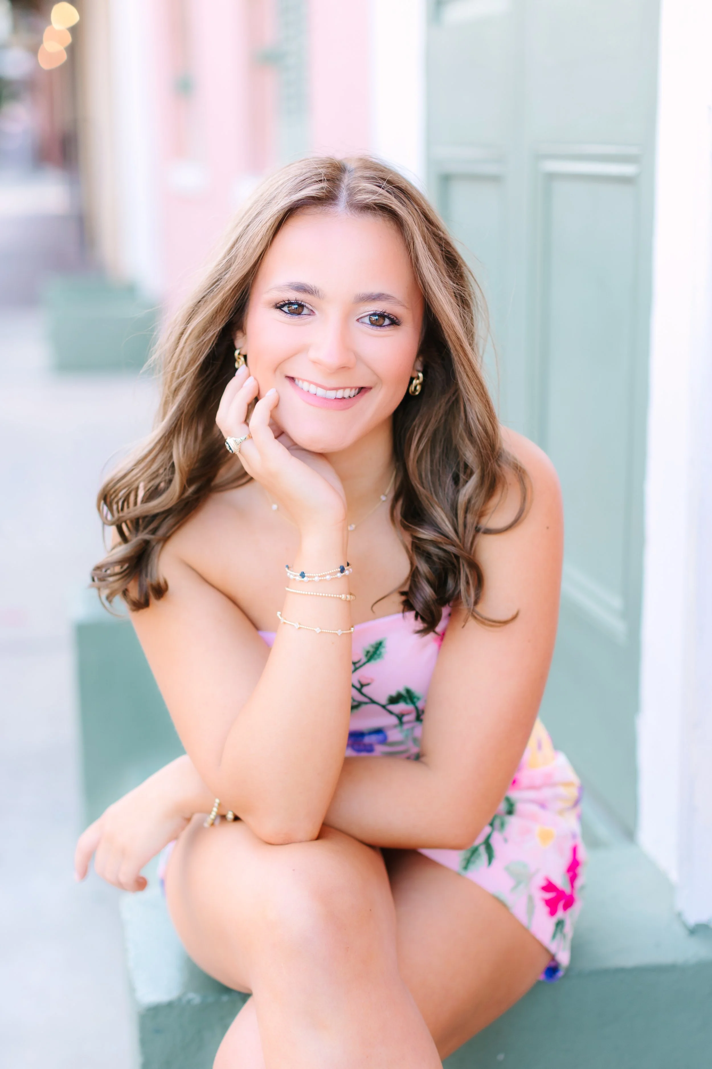 Close-up of a young woman with long brown hair smiling, wearing jewelry, seated outdoors against a pastel-colored building.