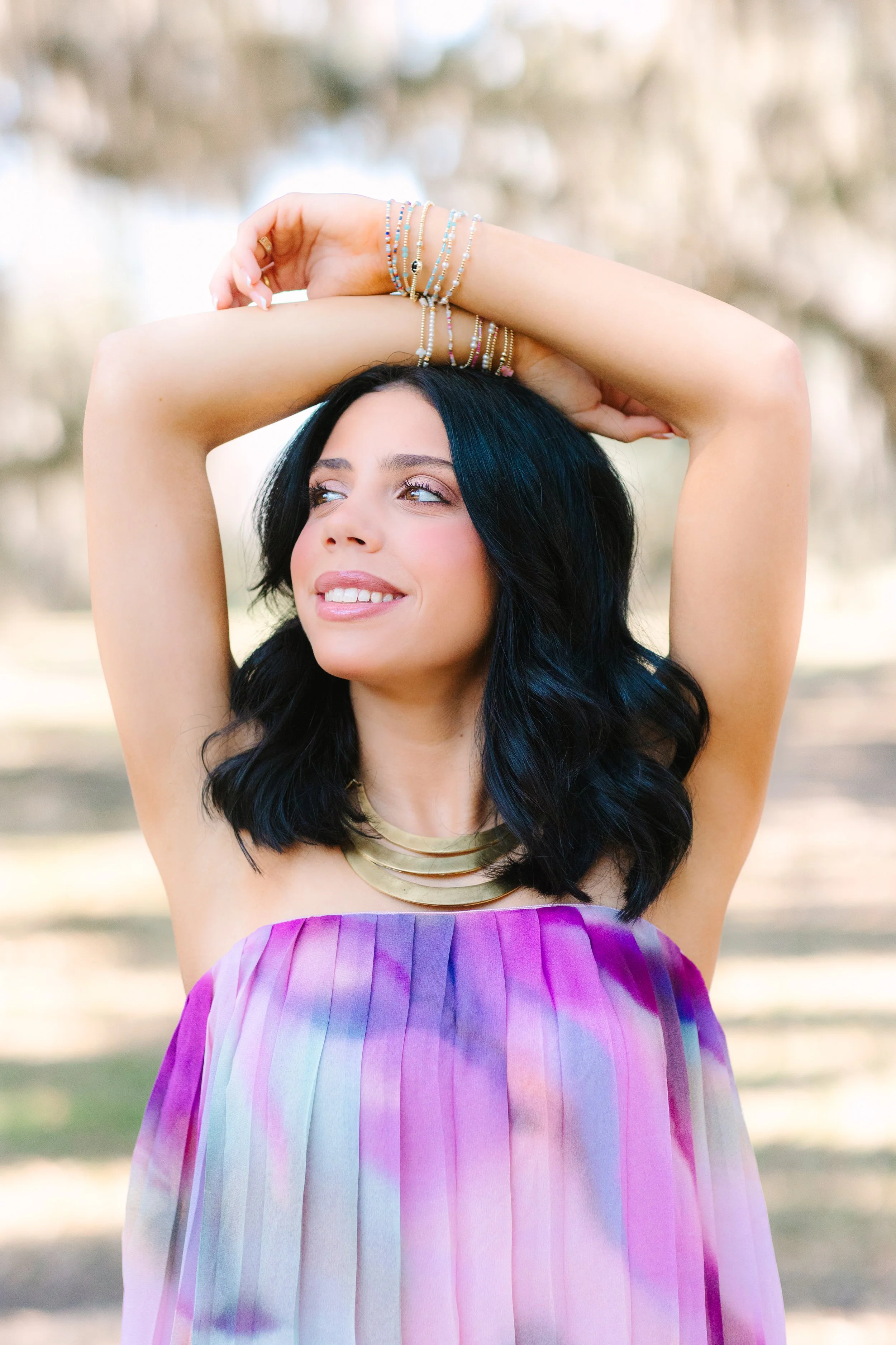A woman has long black hair and tattoos. She wears multiple bracelets, a gold necklace, and a colorful strapless dress with pink and purple hues. She is outdoors with blurred trees in the background and has a serene expression.