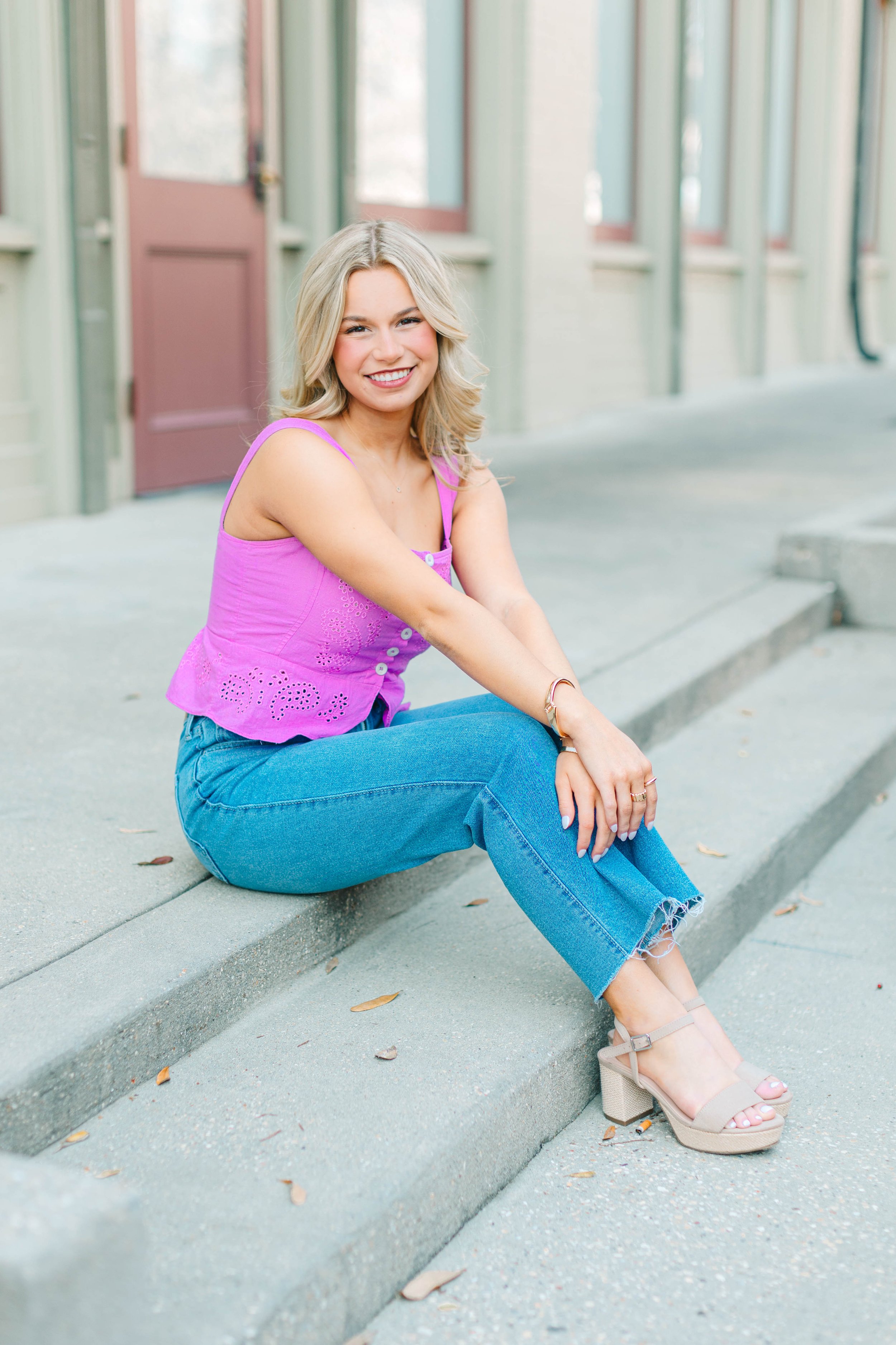 A young woman with blonde hair and light skin is sitting on the sidewalk with her legs crossed and smiling. She is wearing a pink sleeveless top, blue jeans, and beige platform sandals. The background features a building with large windows and a pink door.