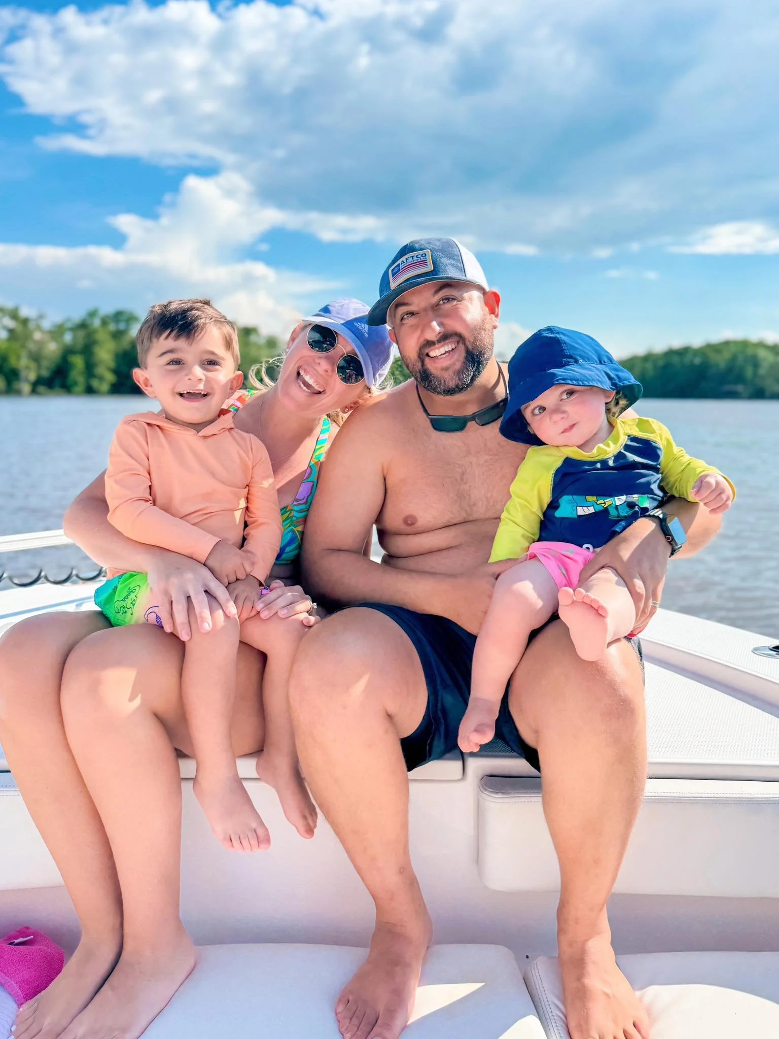 A happy family of four on a boat, with a lake and trees in the background. The woman and man are sitting with their two children, all smiling and enjoying a sunny day.