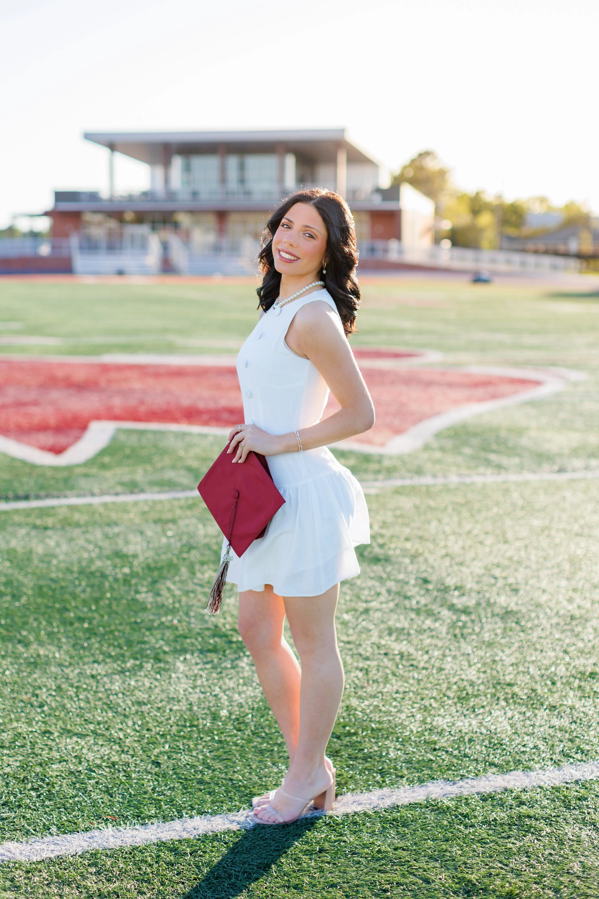 A woman in a white dress holding a maroon graduation cap, standing on a track field at sunset, smiling and looking at the camera.