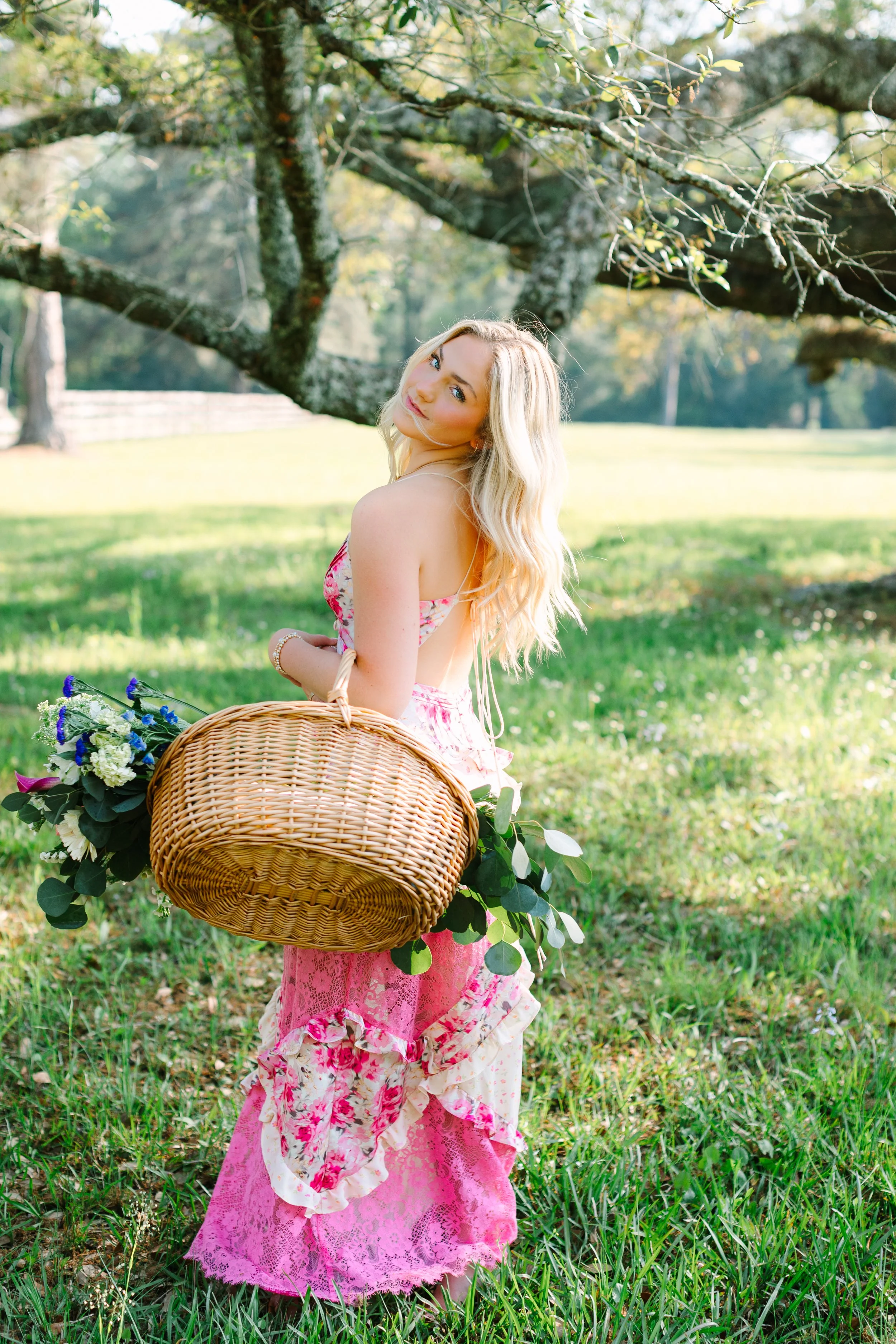 A young woman with long blonde hair wearing a pink floral dress with ruffled skirt, standing outdoors on grass under a large tree with sprawling branches. She is holding a wicker basket decorated with green leaves and white and purple flowers, and is looking back over her shoulder with a gentle expression on her face.