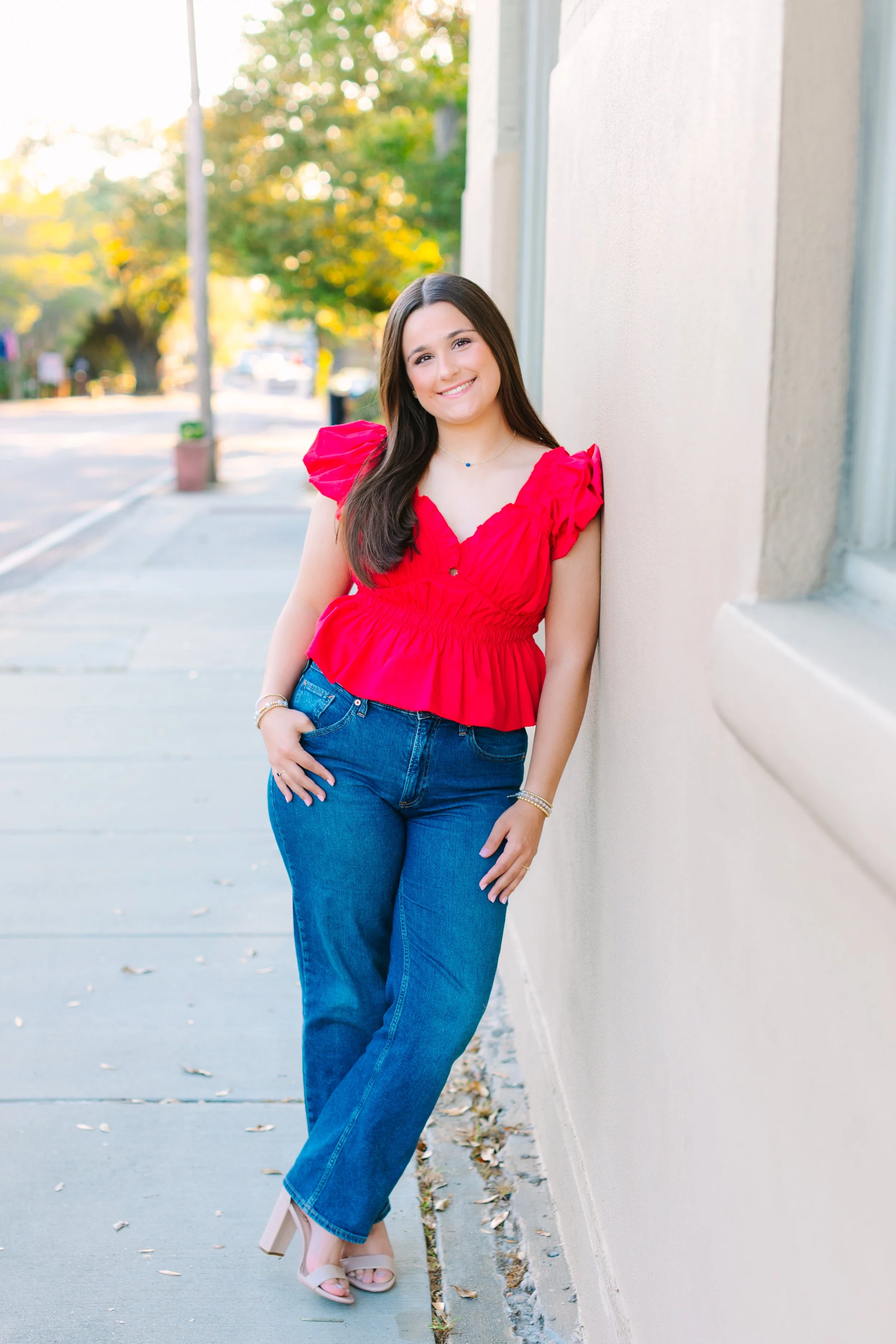 Young woman with long dark hair, wearing a red top with ruffled sleeves, blue jeans, and beige heels, standing outdoors on a sidewalk next to a light-colored wall, smiling at the camera with trees and cars in the background.