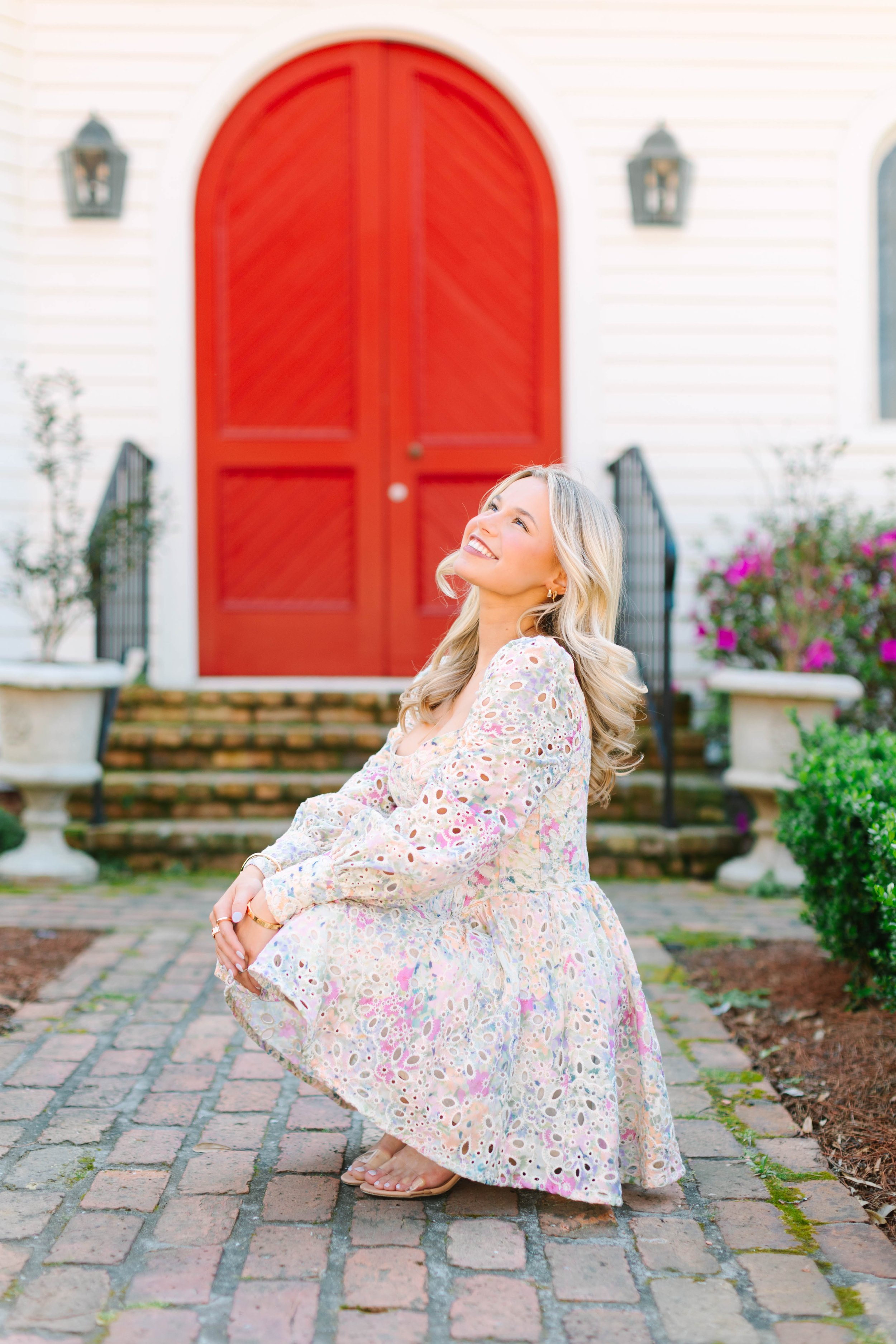 A woman with blonde hair in a floral dress smiling and sitting on her legs on a cobblestone sidewalk in front of a house with white siding and large red double doors. There are potted plants and flowers nearby.