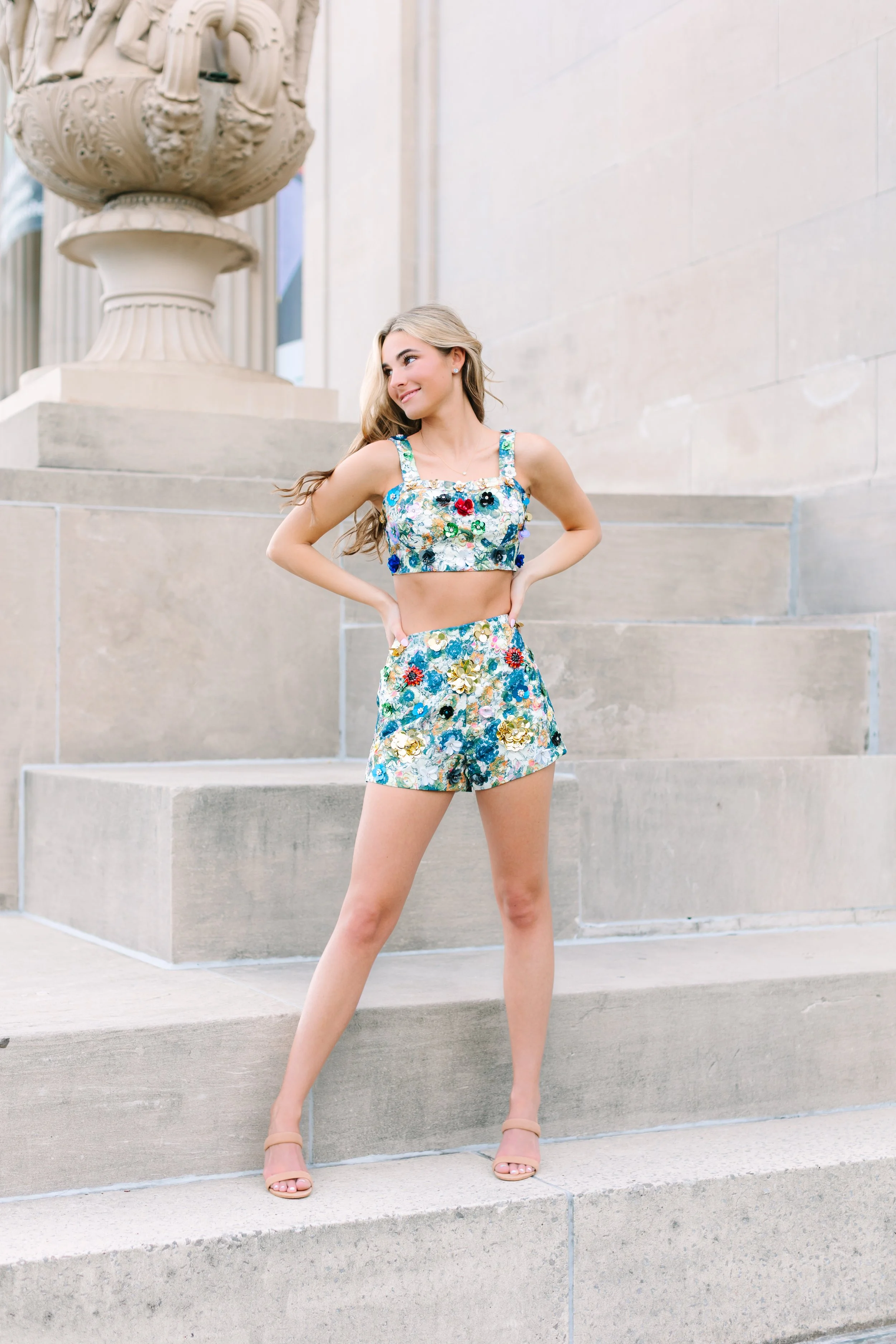 A young woman in a floral crop top and matching shorts posing outside on stone steps with a large decorative urn in the background.