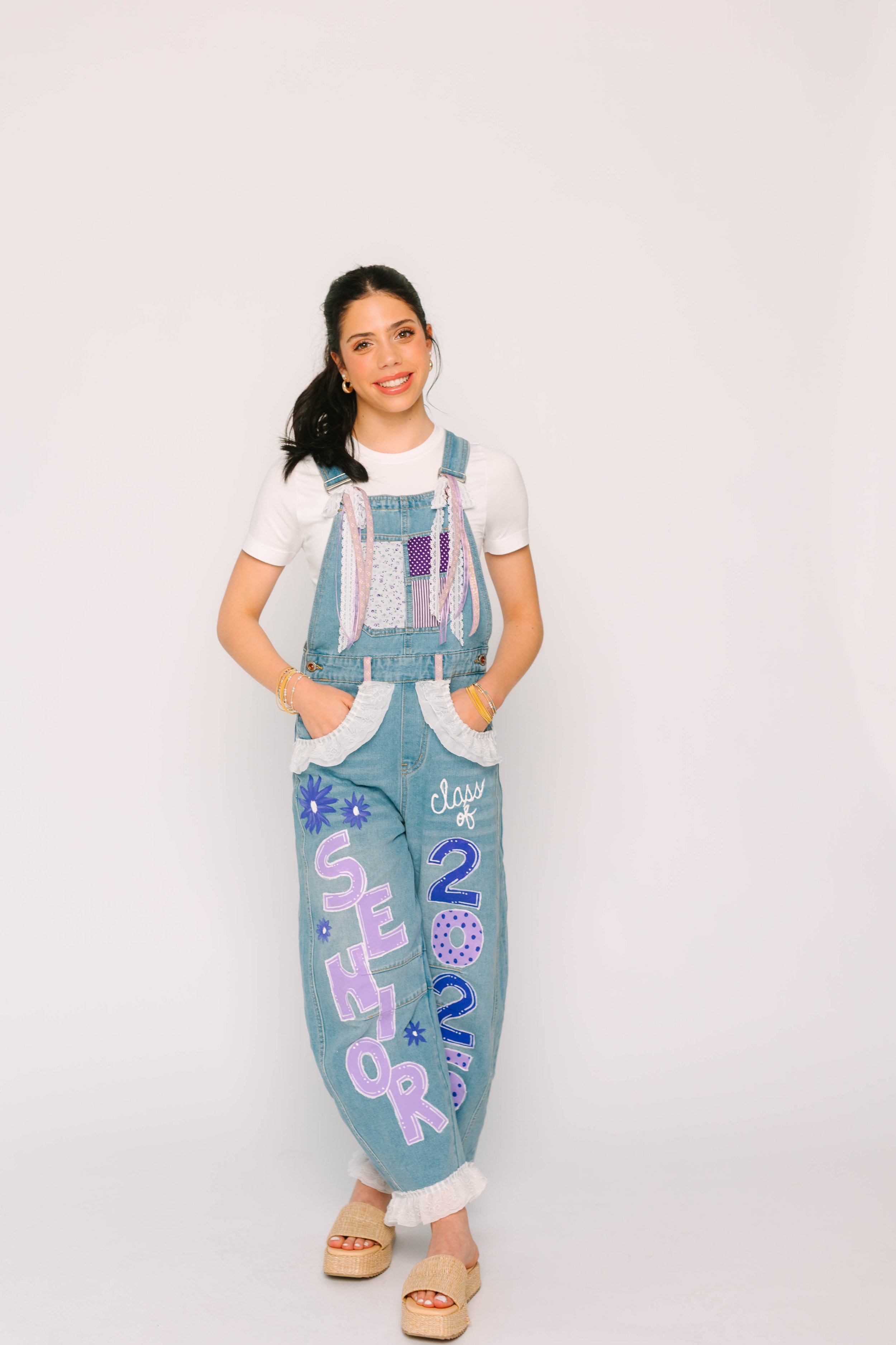 Young woman with dark hair in a ponytail, smiling, wearing a white t-shirt, denim overalls with colorful patches and text, and tan platform slide sandals, standing against a plain white background.