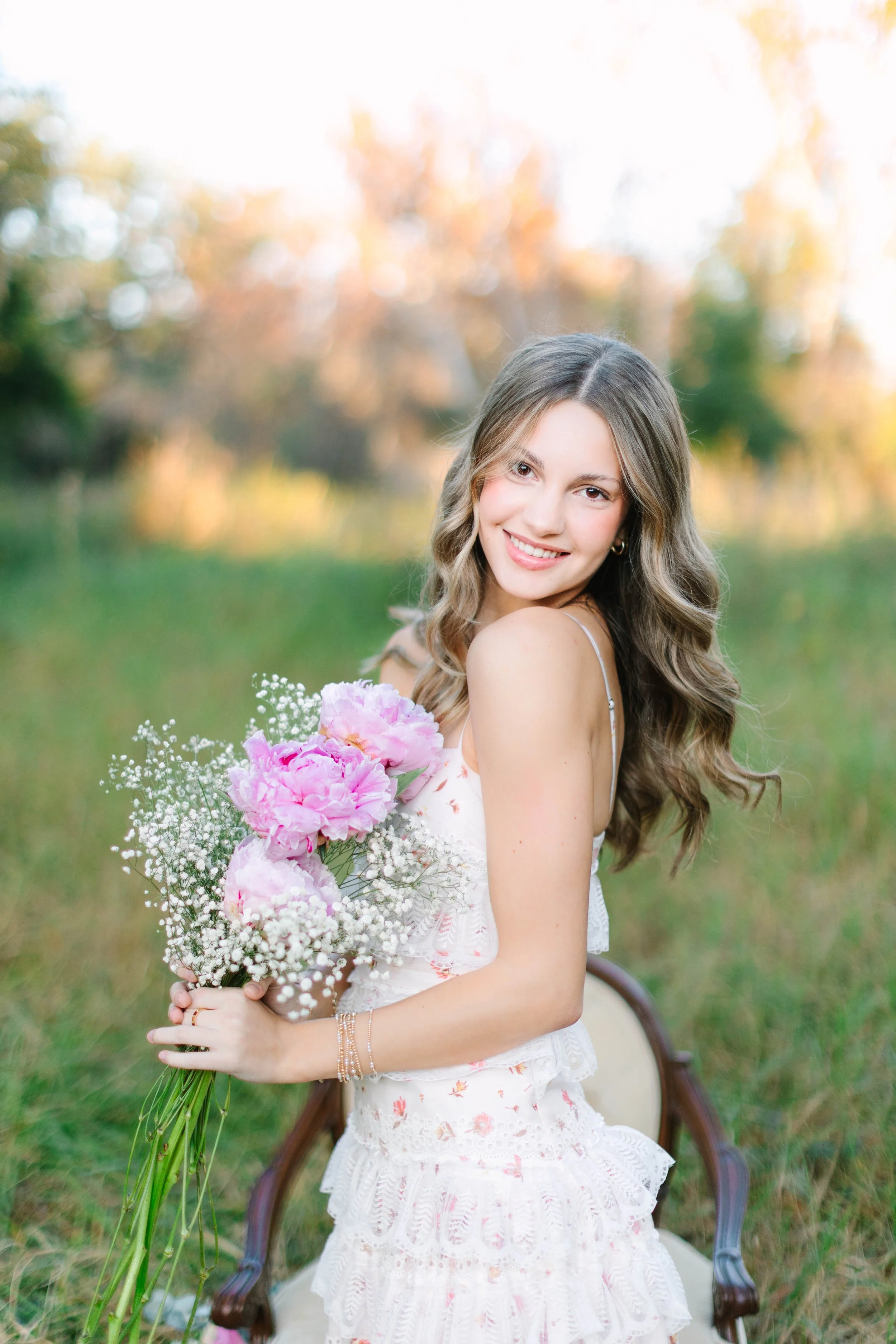 A young woman with long, wavy hair smiling and holding a bouquet of pink and white flowers, sitting outdoors on a wooden chair in a grassy area with blurred trees and sunset in the background.