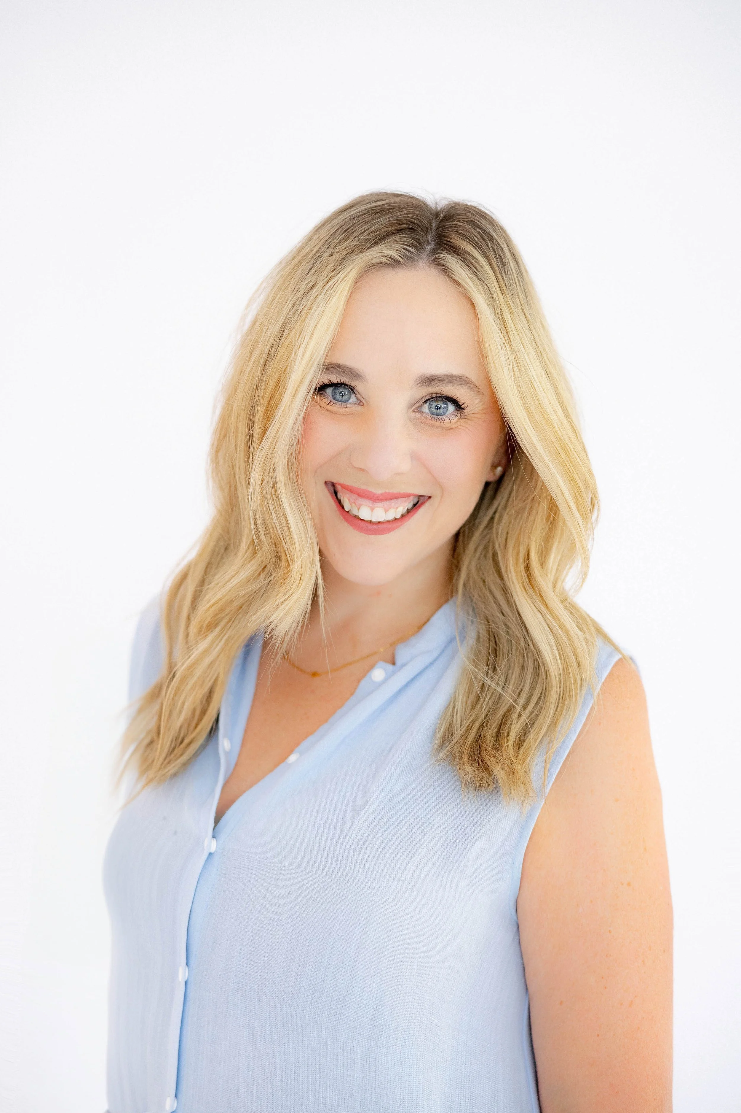 A young woman with long blonde hair and blue eyes smiling, wearing a sleeveless light blue collared top, against a plain white background.