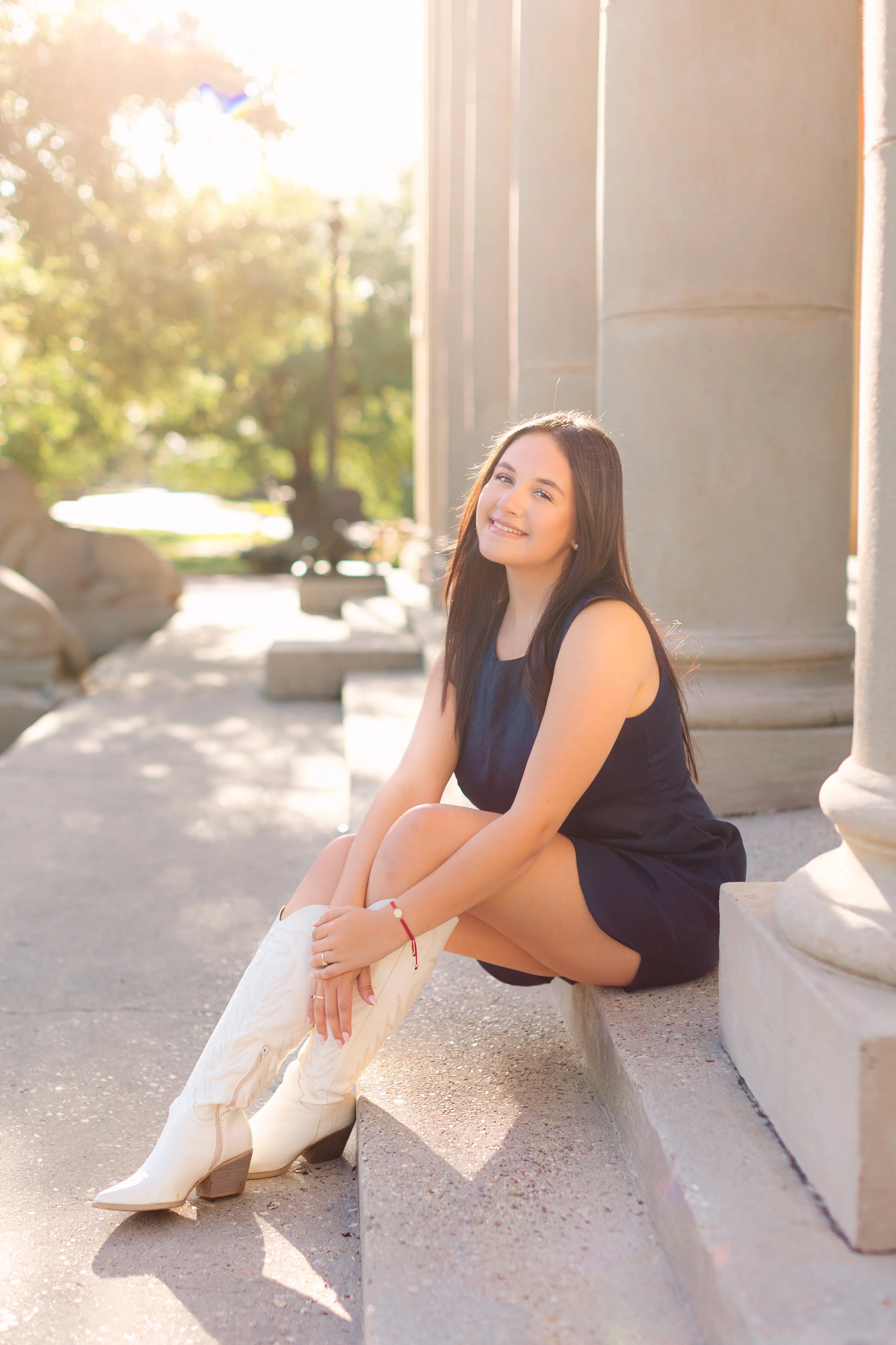 A young woman with long brown hair sitting on a stone bench outside during sunset, wearing a dark sleeveless dress, white boots, and a red bracelet, smiling at the camera.