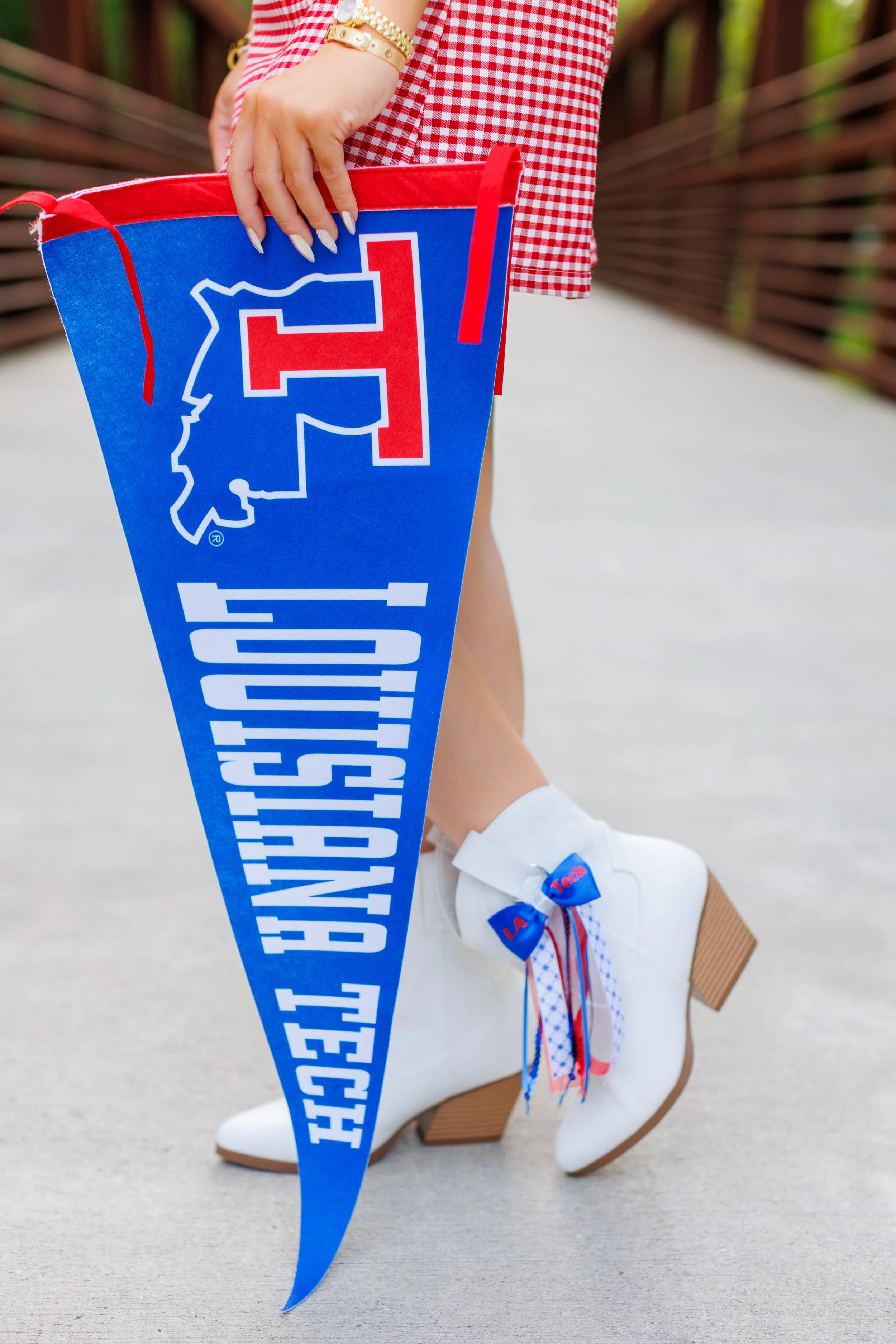 Close-up of a person holding a large blue 2023 graduation cap with the outline of a state and the year 2023 on it. The person is wearing a red and white checkered sleeve, gold bracelets, white shoes with blue and red ribbons, white socks, and is on a bridge or walkway with a blurred background of greenery.