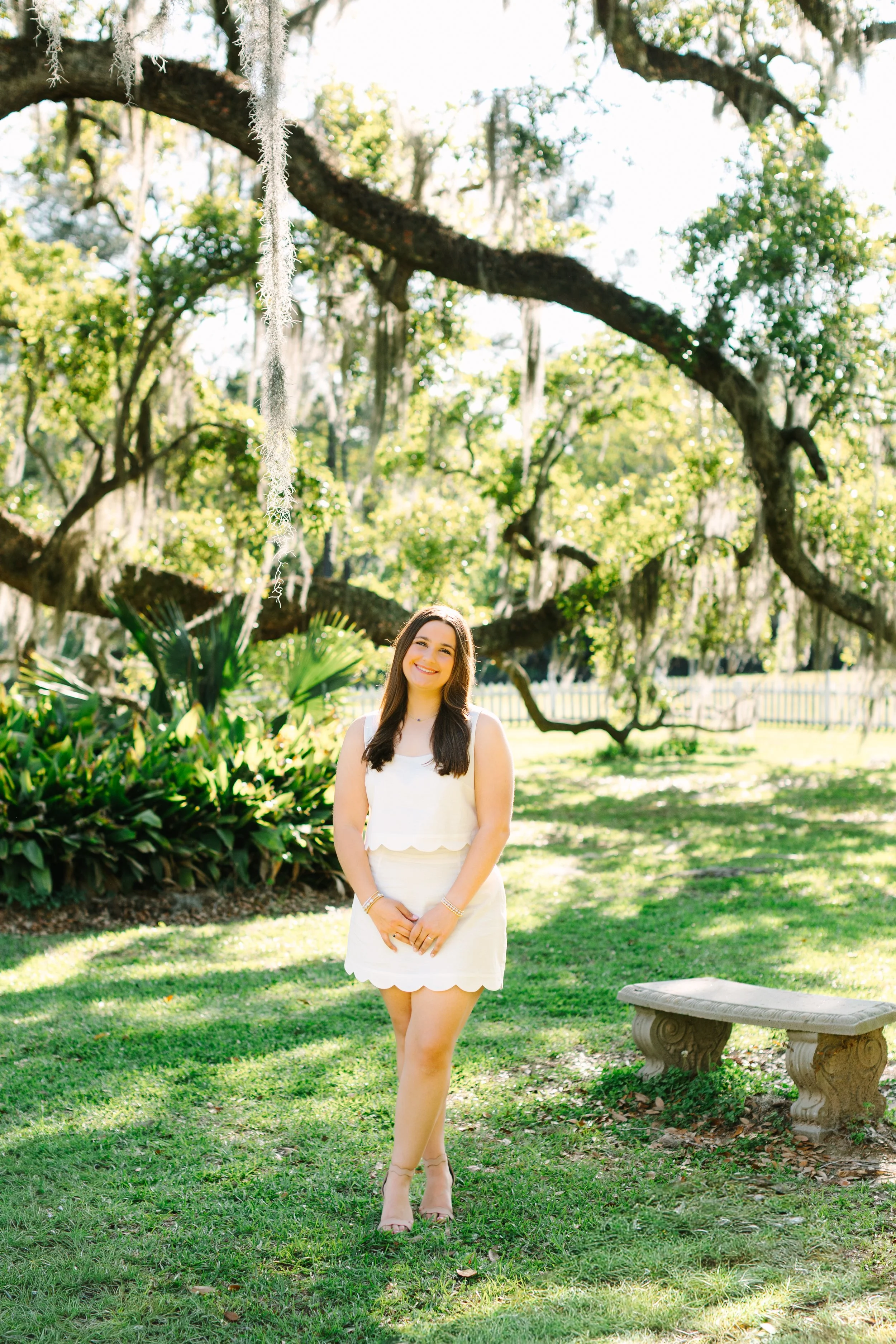 A woman standing outdoors on a grassy lawn, smiling, with trees and a stone bench in the background.