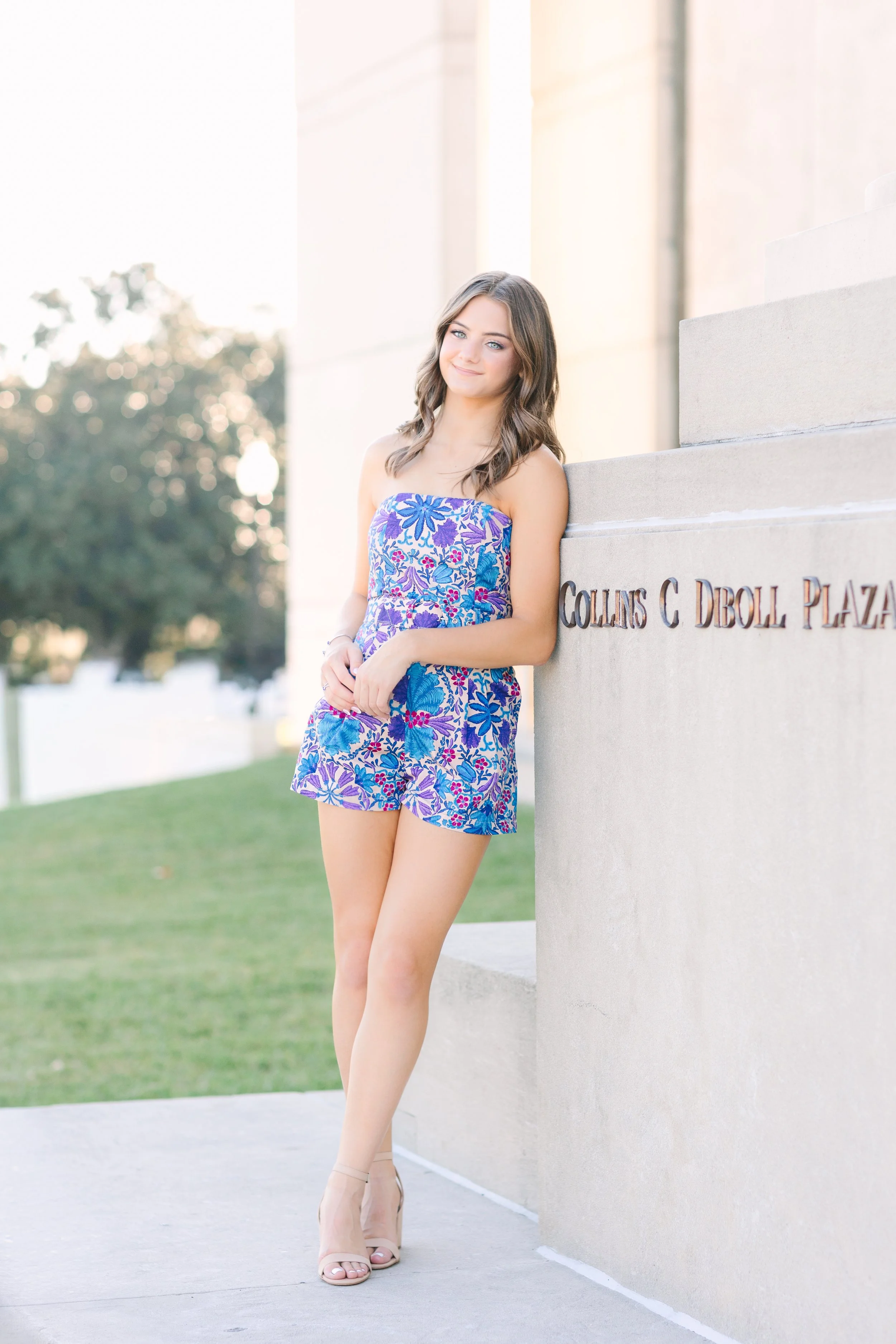 A young woman with shoulder-length brown hair and fair skin stands smiling outdoors next to a stone wall with the text "Collins C. Droll Plaza". She wears a strapless, colorful floral romper and high heels, with a background of trees and a grassy area.