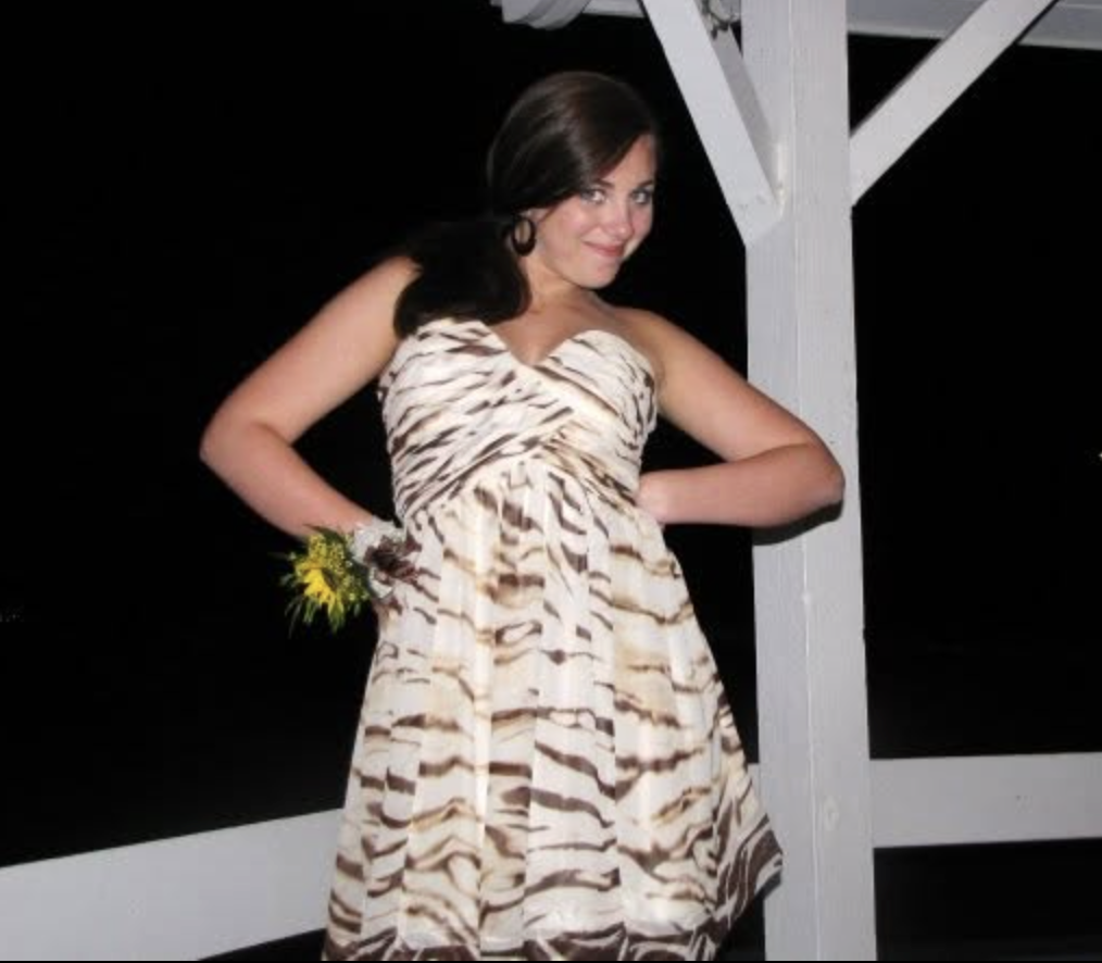 A woman with dark hair and earrings wearing a zebra-print dress, standing outdoors at night against a white wooden structure, smiling and posing with her hands on her hips.