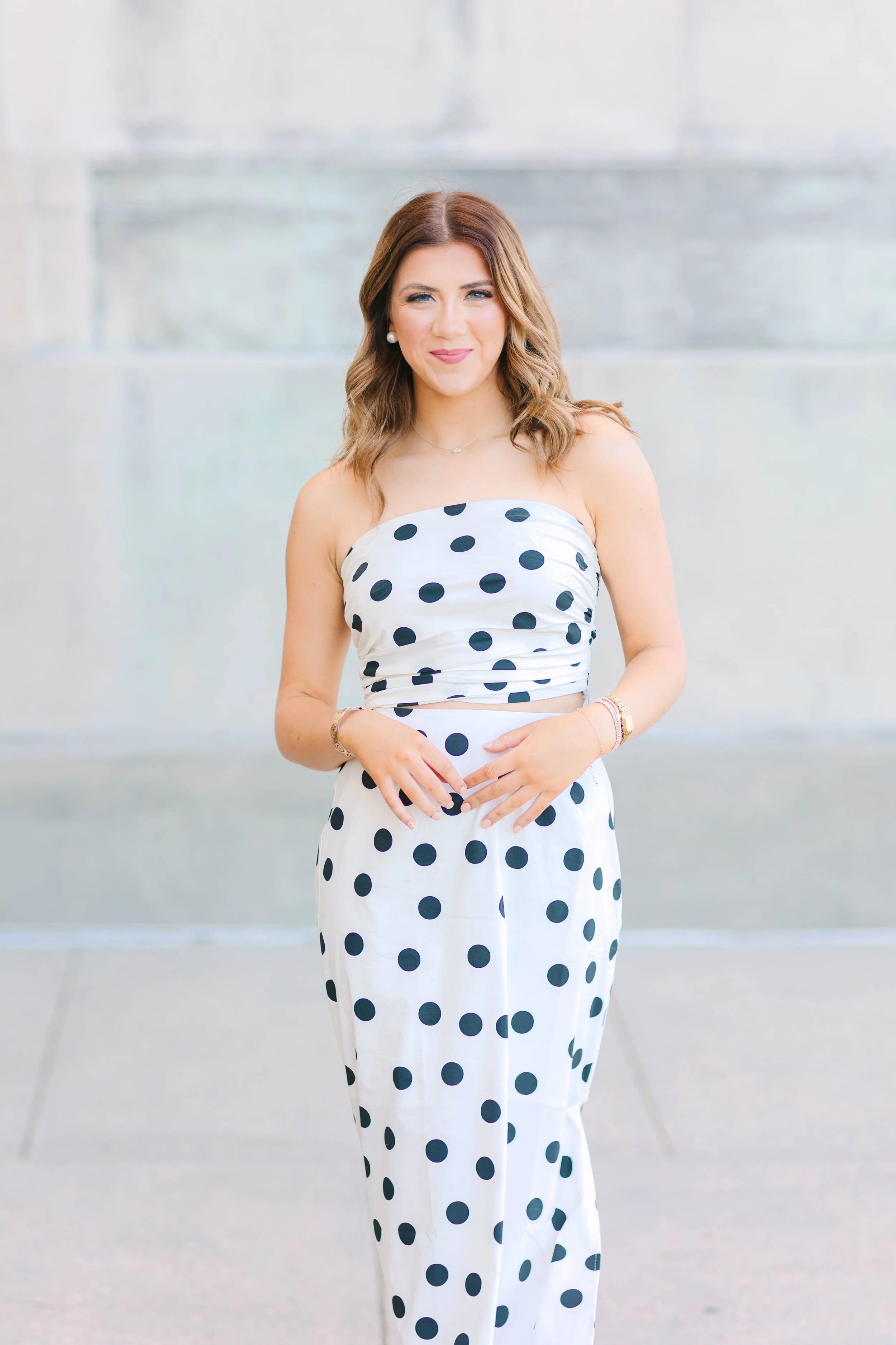 A woman with shoulder-length wavy brown hair, wearing a strapless white dress with black polka dots, standing with her hands in front of her against a blurred gray background.
