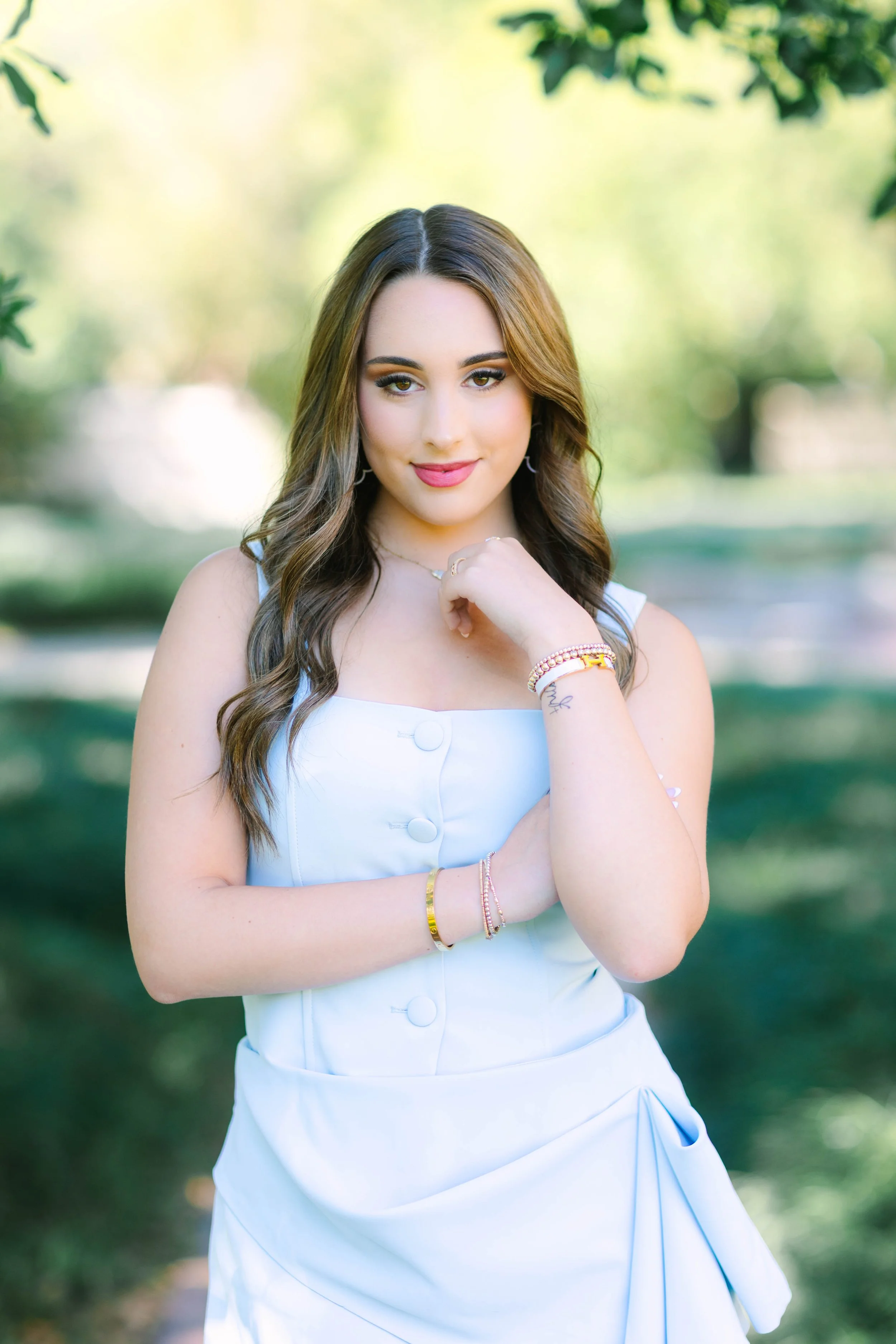 Young woman with brown hair wearing a white sleeveless dress, jewelry, and makeup outdoors with green trees blurred in the background.