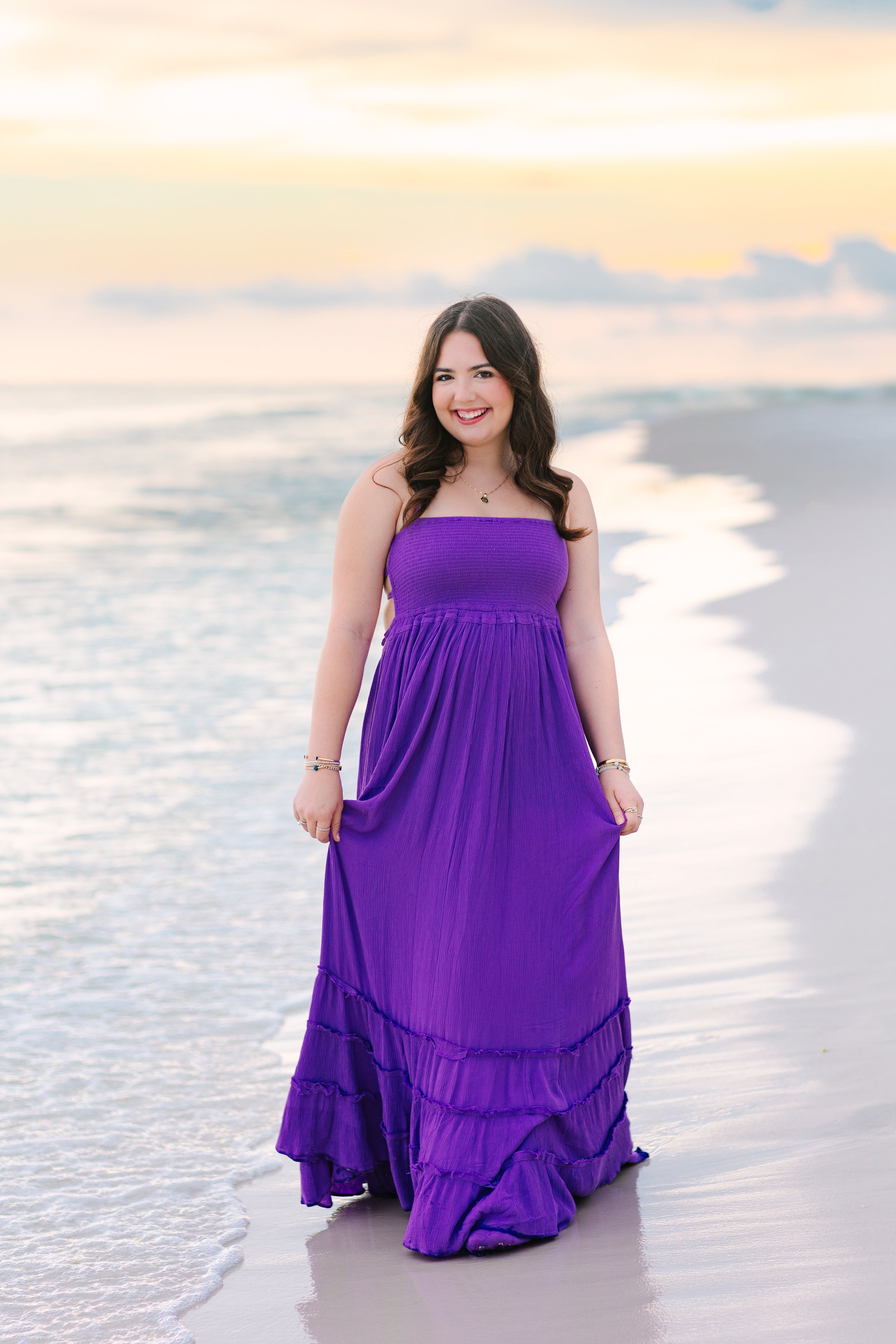 A woman smiling at the camera, wearing a purple strapless dress, standing on wet sand by the ocean at sunset.