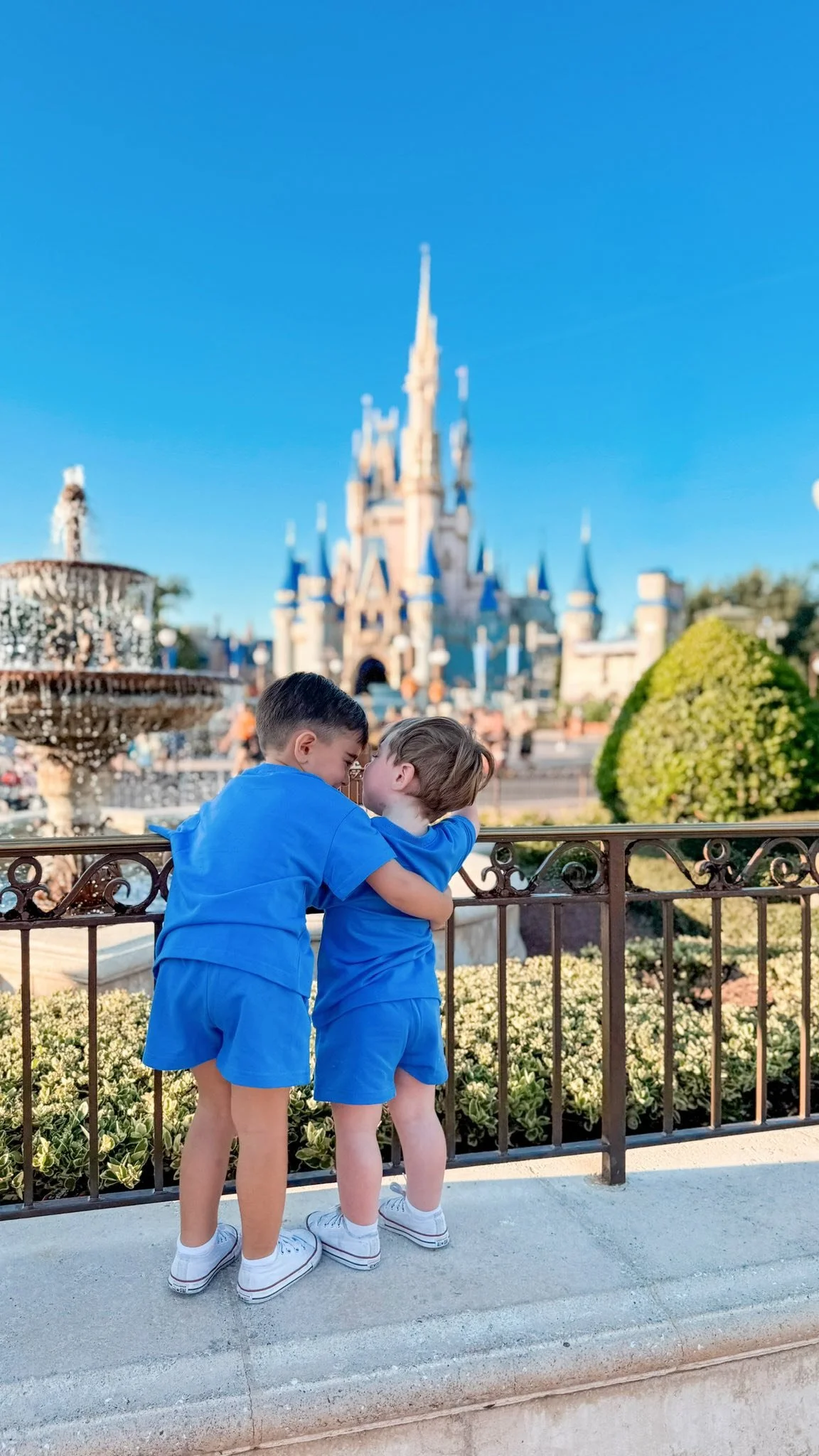Two young boys in blue shorts and shirts share a kiss at a theme park with a castle, fountain, and landscaping in the background.