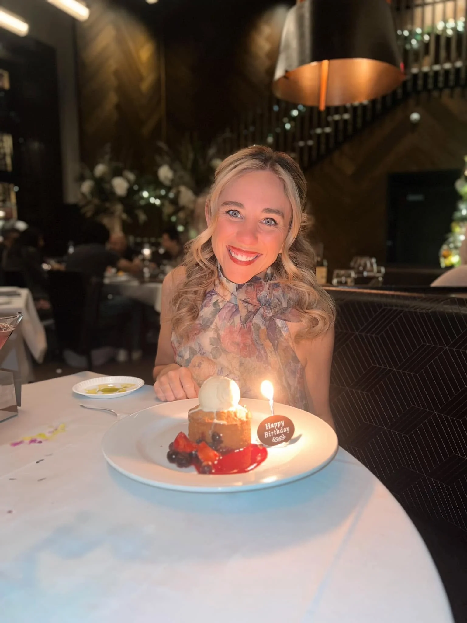 A woman with blonde curly hair smiling at a birthday celebration, sitting in front of a dessert with a lit candle, in a dimly lit restaurant.