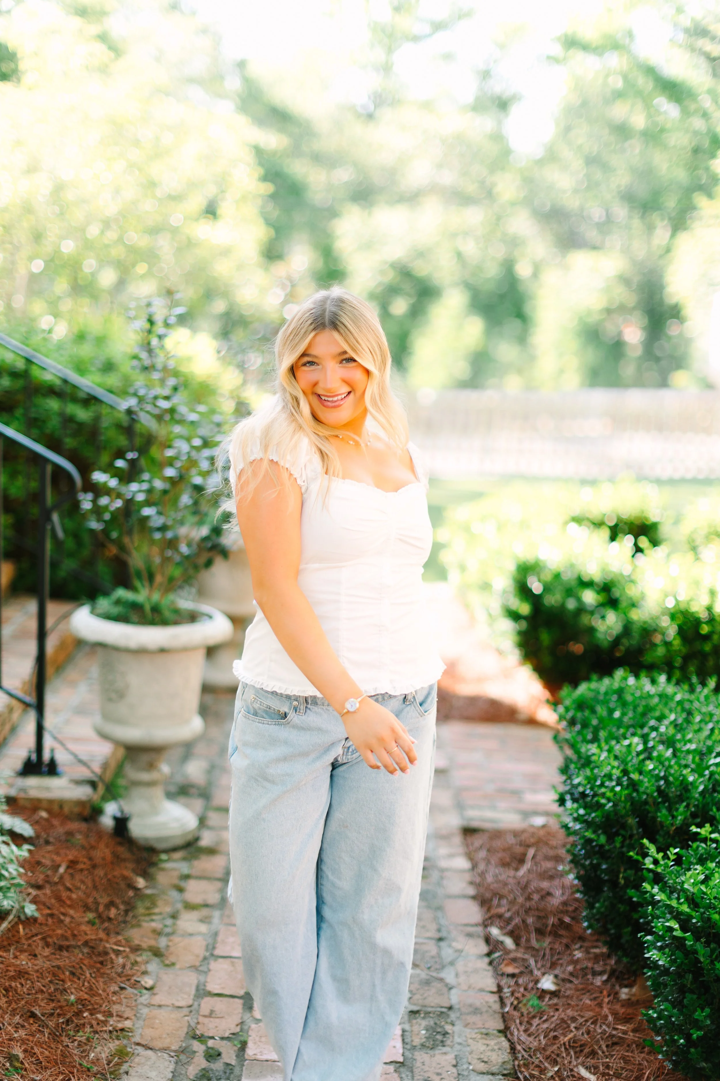 A woman with blonde hair wearing a white top and light blue jeans standing outdoors on a brick path surrounded by greenery, smiling at the camera.