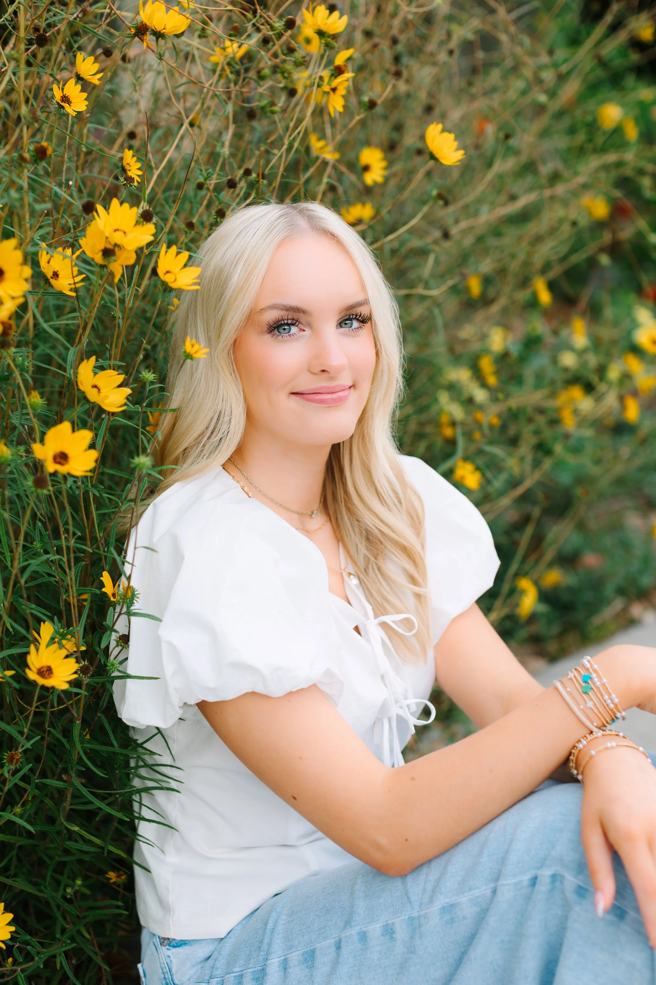 A young woman with blonde hair and blue eyes sitting outdoors among yellow flowers, wearing a white top and light blue jeans, smiling softly at the camera.