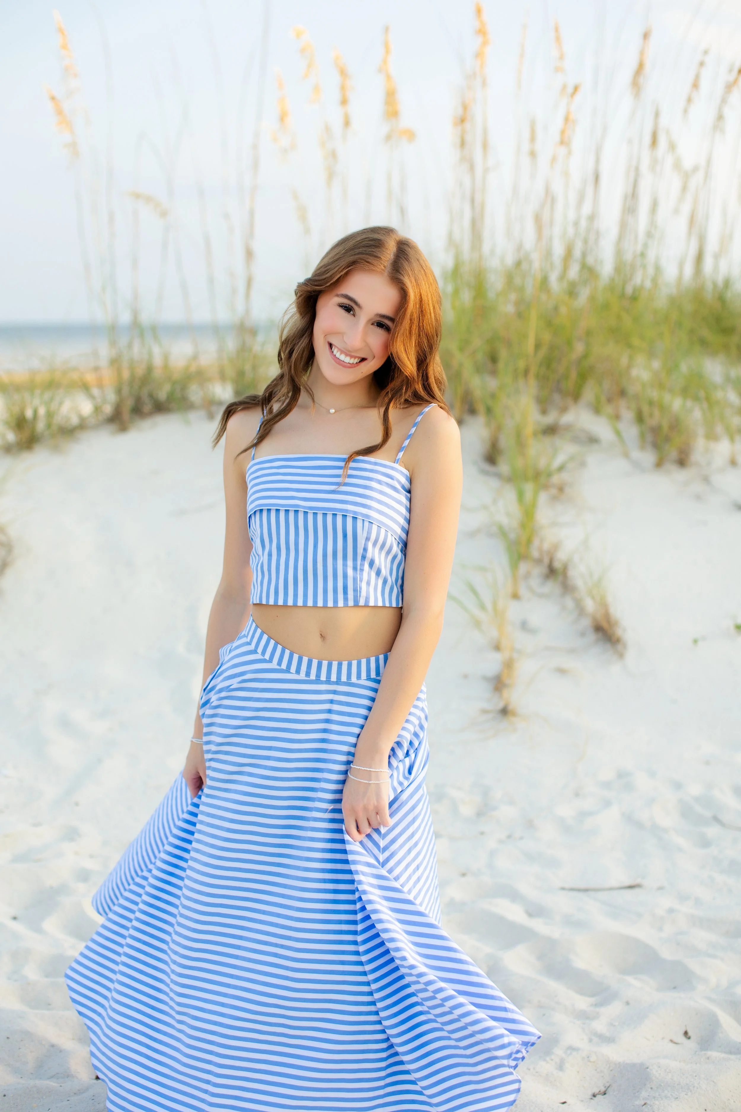 A woman with brown, wavy hair wearing a blue and white striped crop top and matching skirt, smiling on a sandy beach with tall grass in the background.