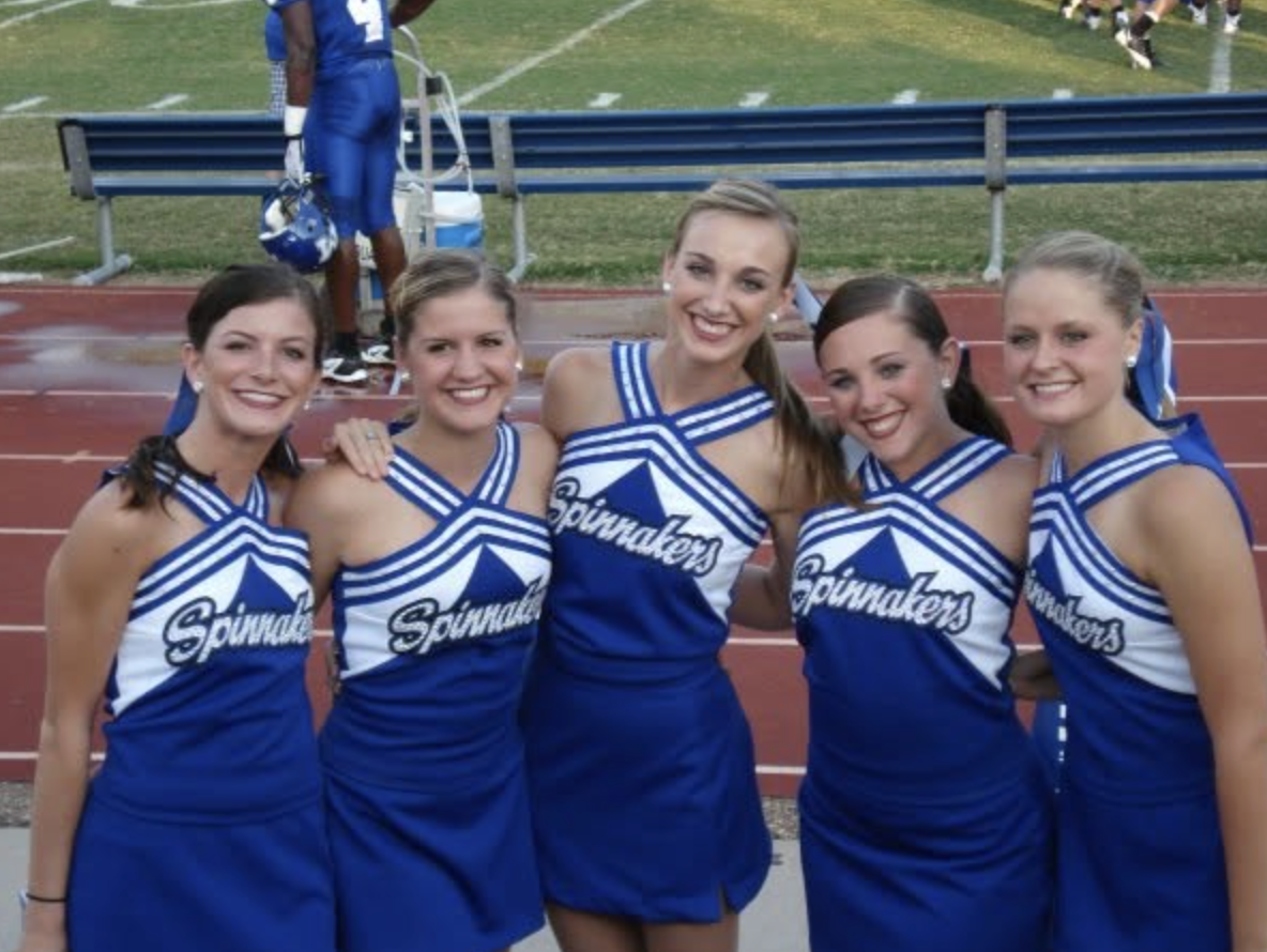 Five cheerleaders in blue and white uniforms with 'Spinnakers' written on them, posing on a track at a sports field, smiling.