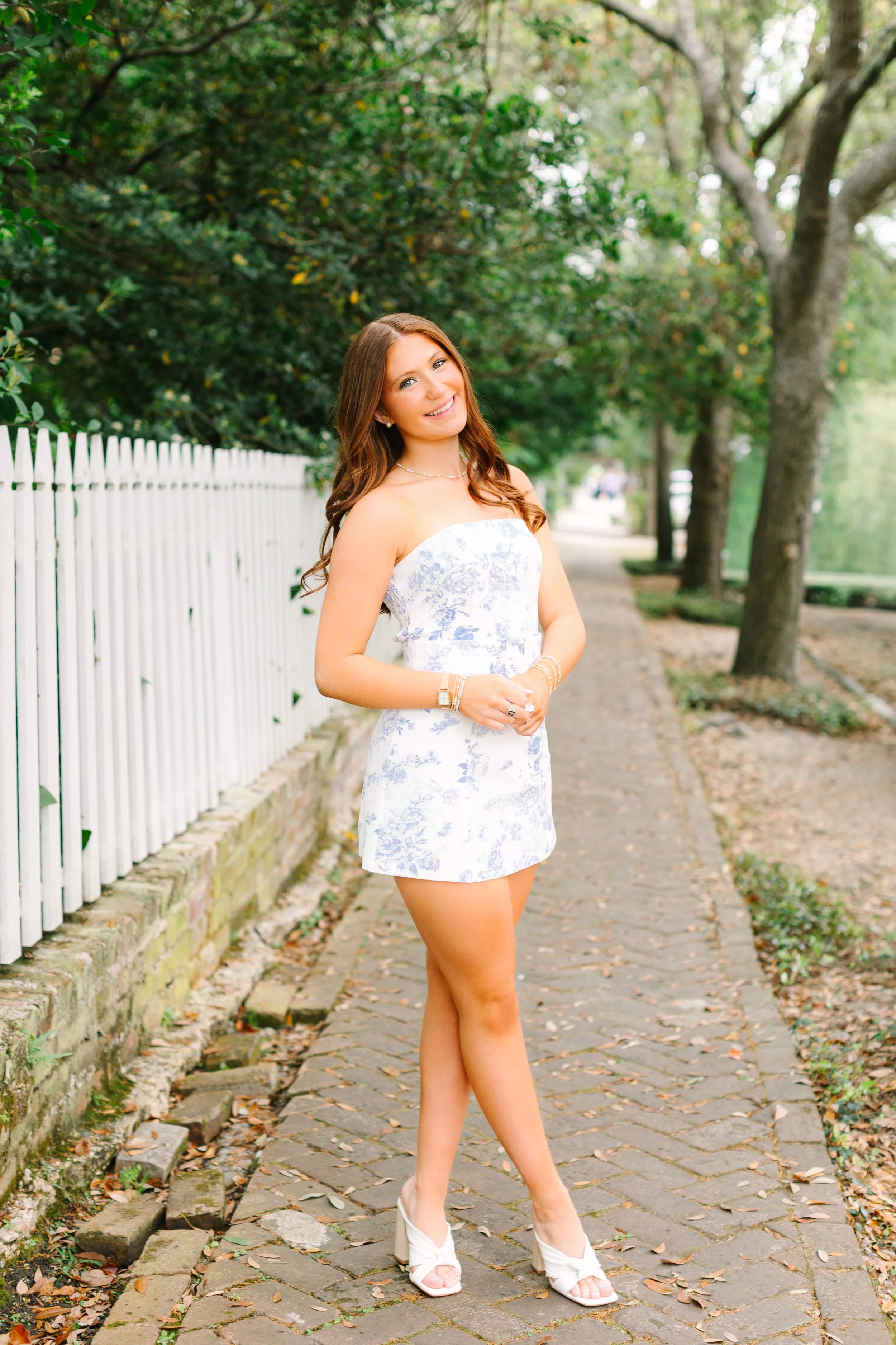 Young woman in white floral dress and white high heels standing on cobblestone path with trees and white picket fence in the background