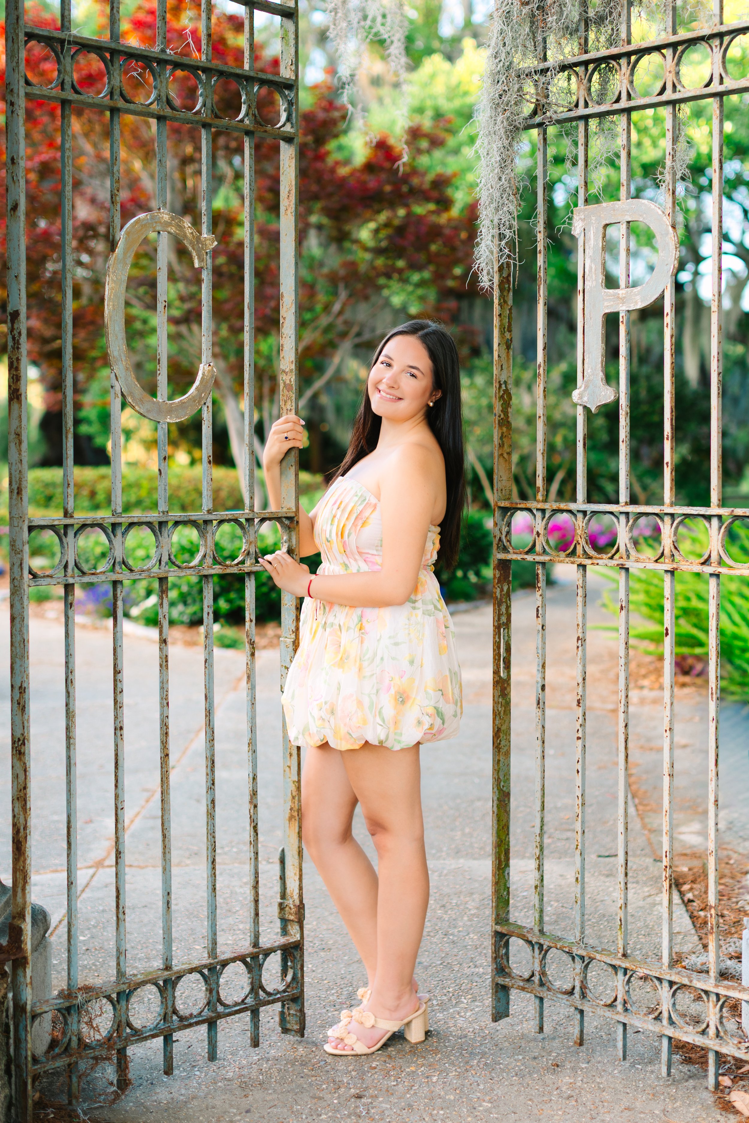 A young woman with long dark hair, wearing a strapless floral dress and beige platform sandals, standing between rusty metal gates in a lush garden with vibrant red and green foliage.