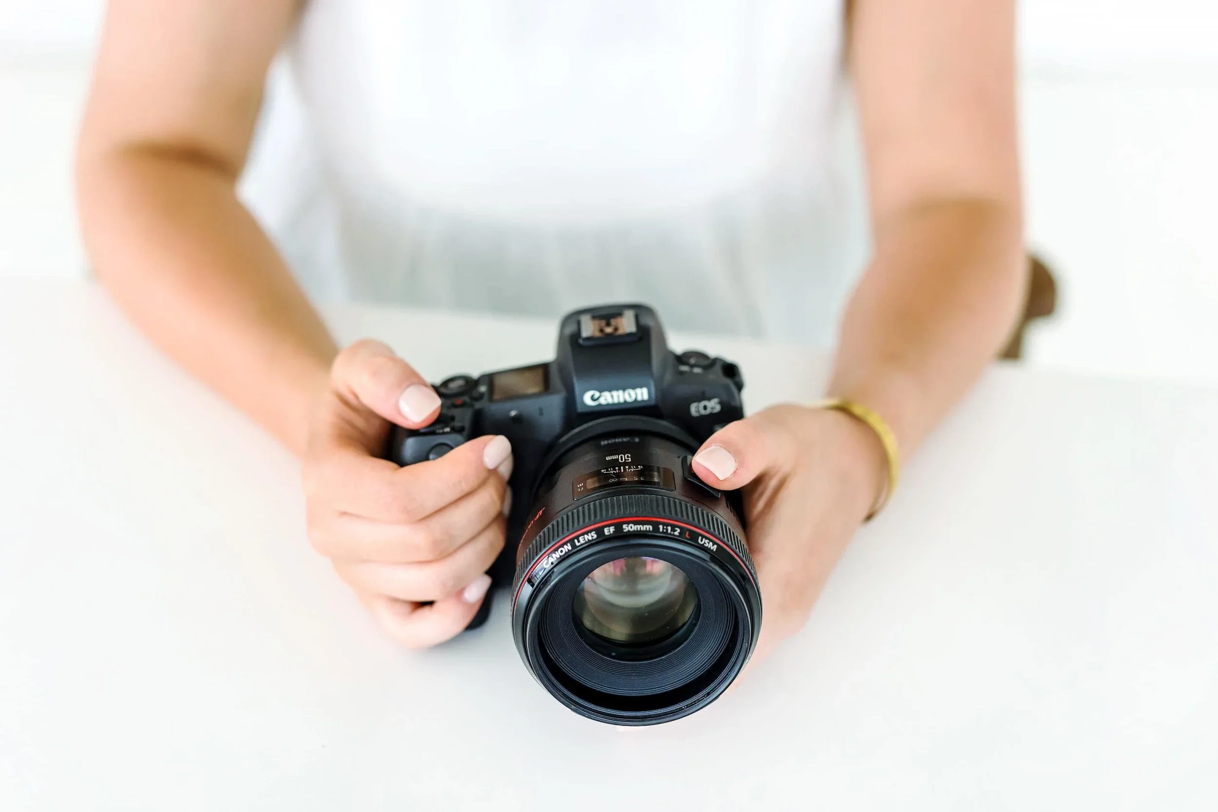 Person holding a Canon DSLR camera with a 50mm lens, wearing a white top, with a white background.