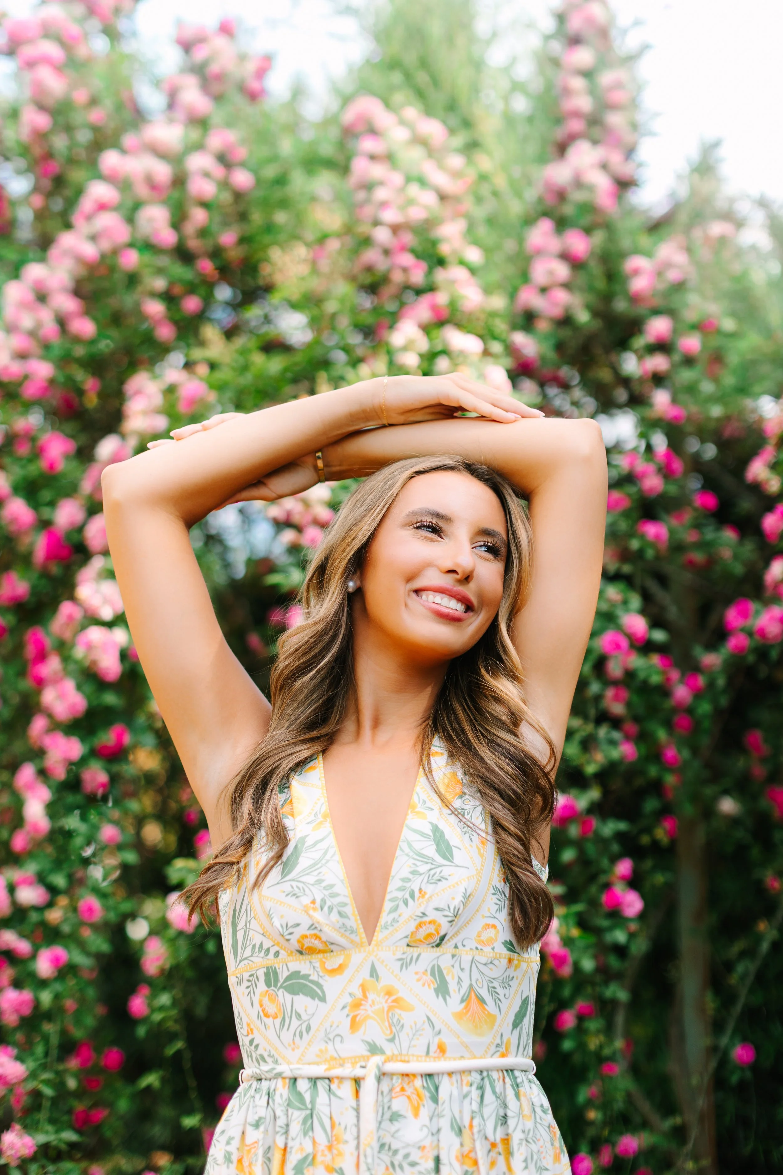 A woman with long wavy hair, smiling, wearing a yellow and white floral dress, standing outdoors in front of a pink flowering bush, with her arms raised above her head.