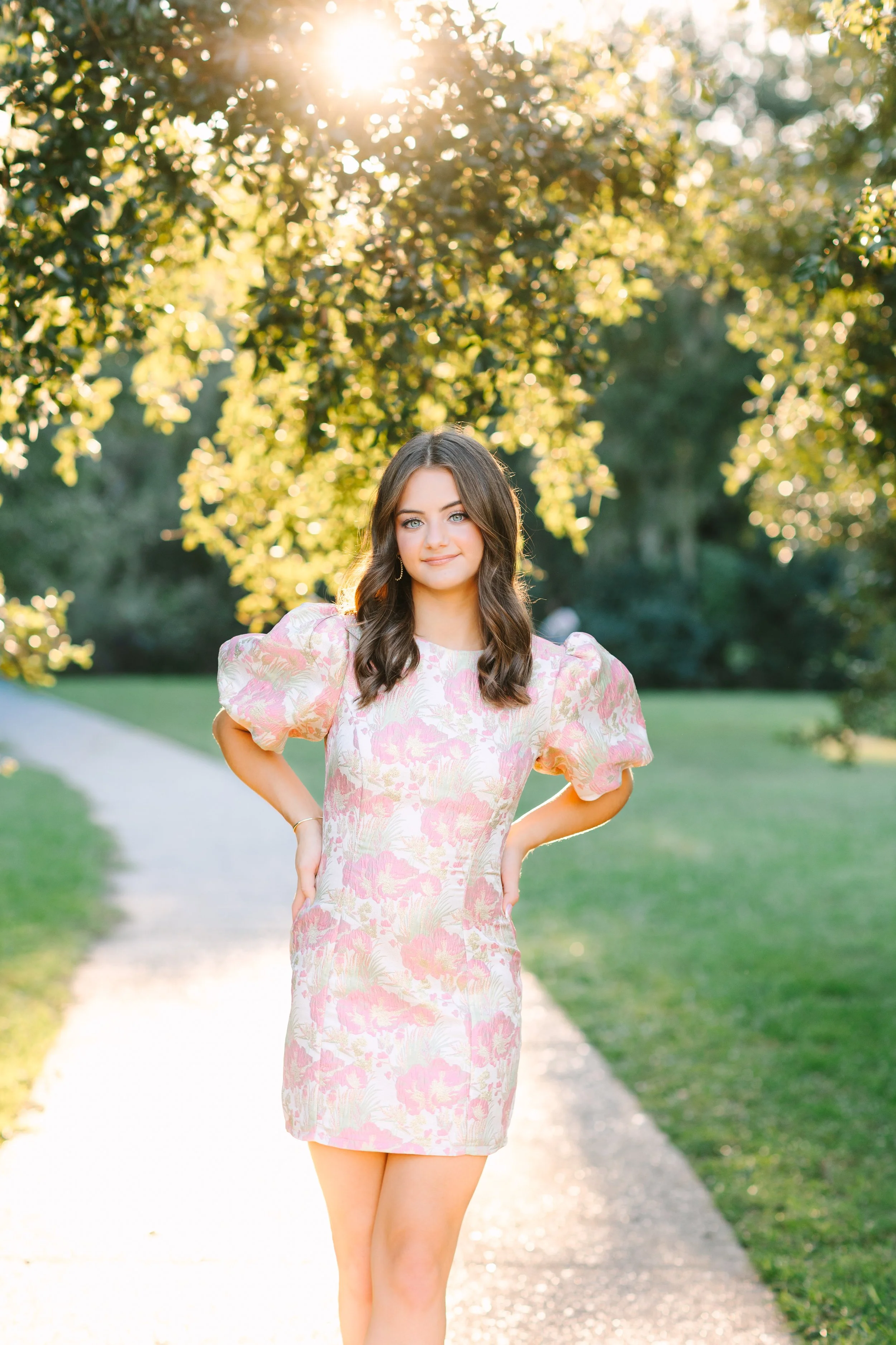 A young woman with long brown hair and blue eyes stands on a sunny park pathway with greenery and trees in the background. She wears a pink and cream floral dress with puffed sleeves and has a confident, relaxed expression.