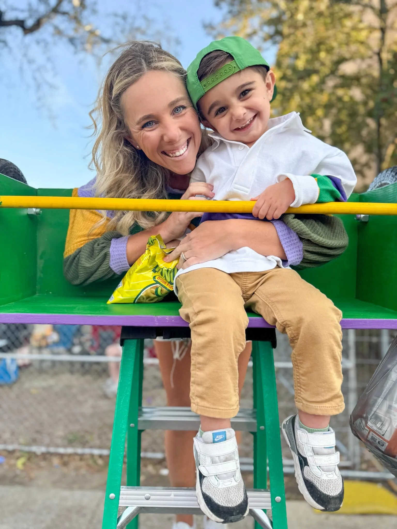 A smiling woman and young boy sitting together on a green and purple amusement park ride. The boy is wearing a green cap and white jacket, and the woman has long blonde hair. They are outdoors with trees in the background, and they look happy.
