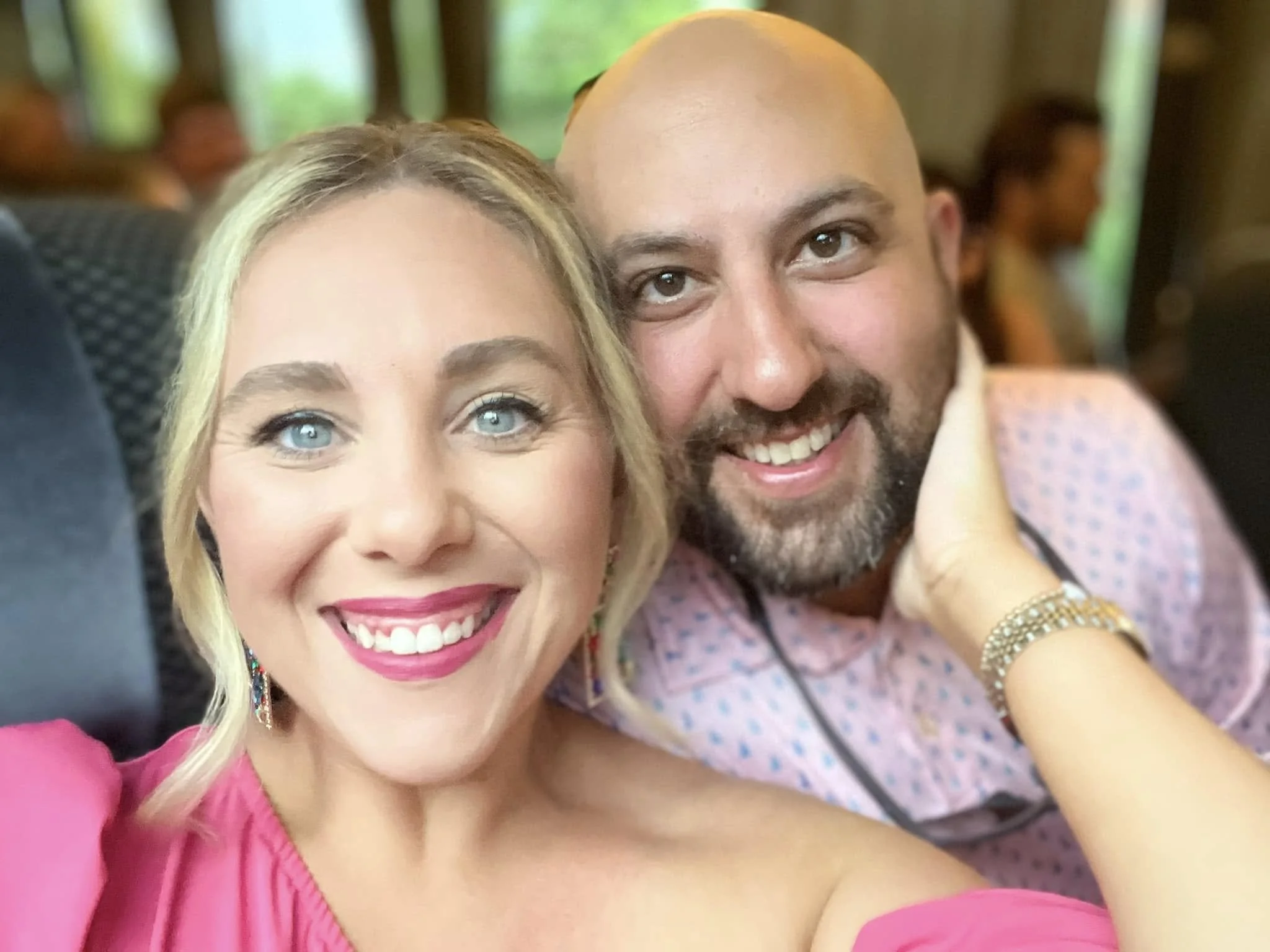 A woman with blonde hair, blue eyes, bright lipstick, wearing earrings and a pink top, smiling next to a man with a bald head, brown eyes, a beard, wearing a patterned shirt, both taking a selfie.