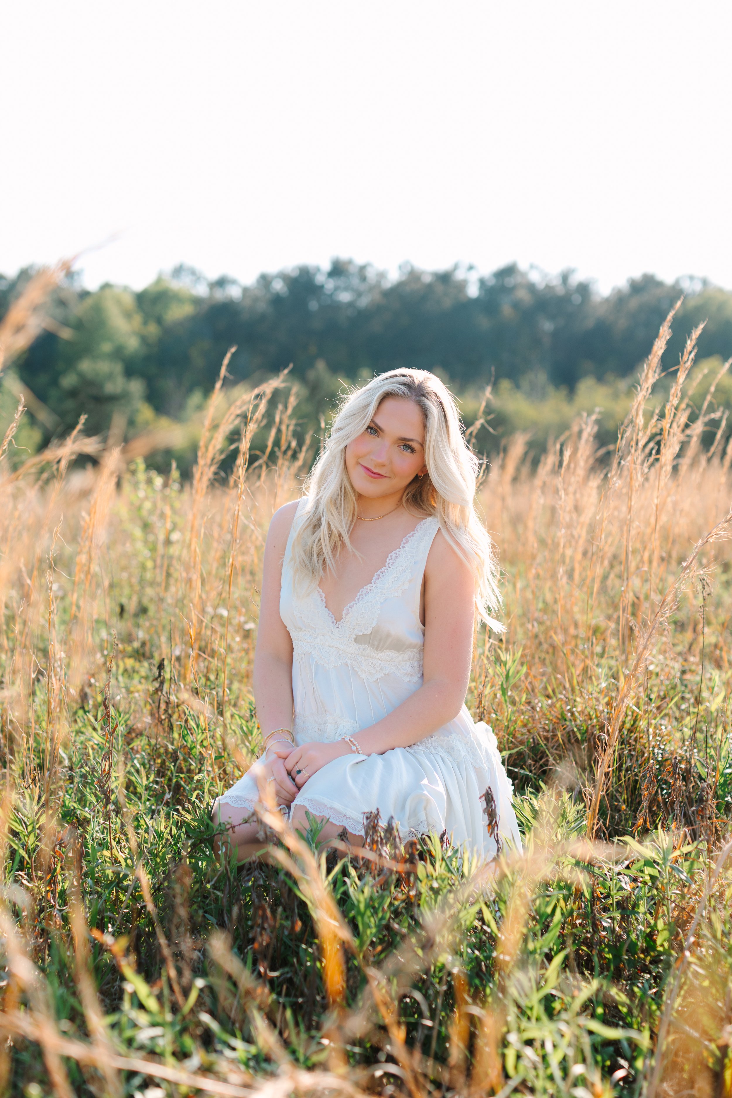 A woman with long blonde hair wearing a white dress sitting in a field of tall grasses with green trees in the background on a sunny day.