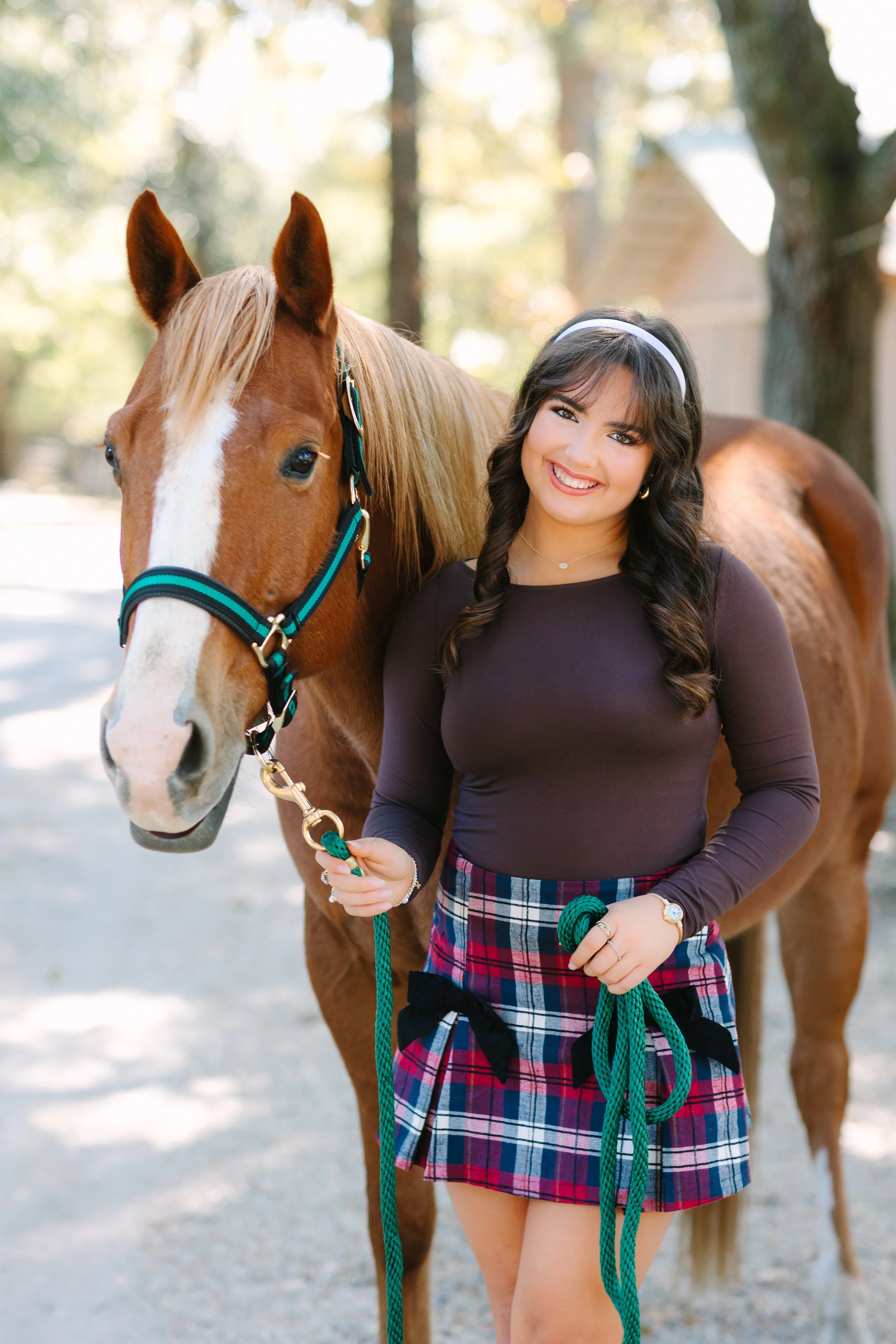 A young woman with long dark hair, wearing a white headband, a dark long sleeve top, and a red plaid skirt, stands outdoors holding a green horse lead rope with a chestnut horse beside her. The background has trees and sunlight filtering through.