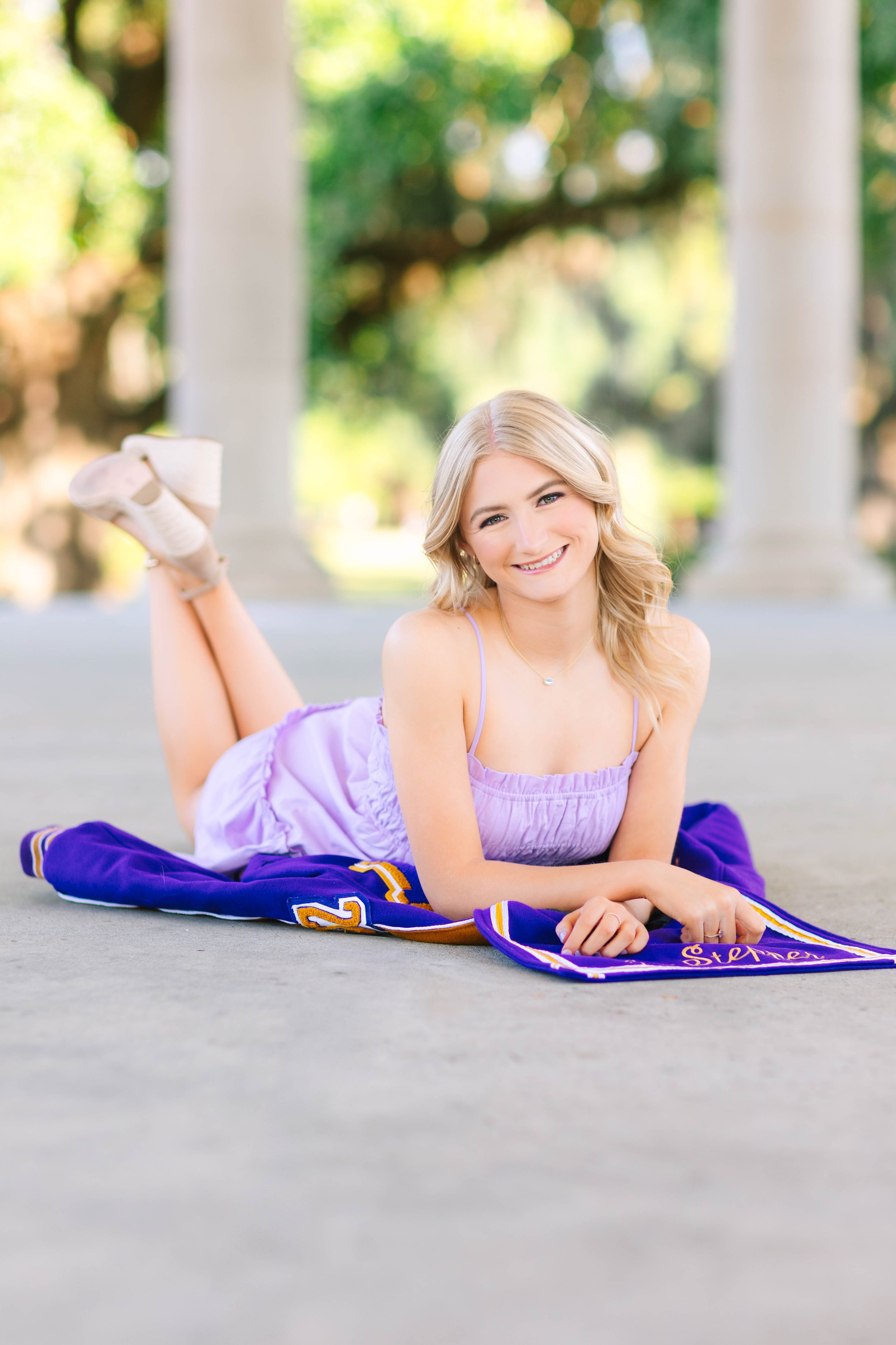 A young woman with blonde hair lying on her stomach on a purple blanket outdoors, smiling at the camera, with trees and greenery in the background.