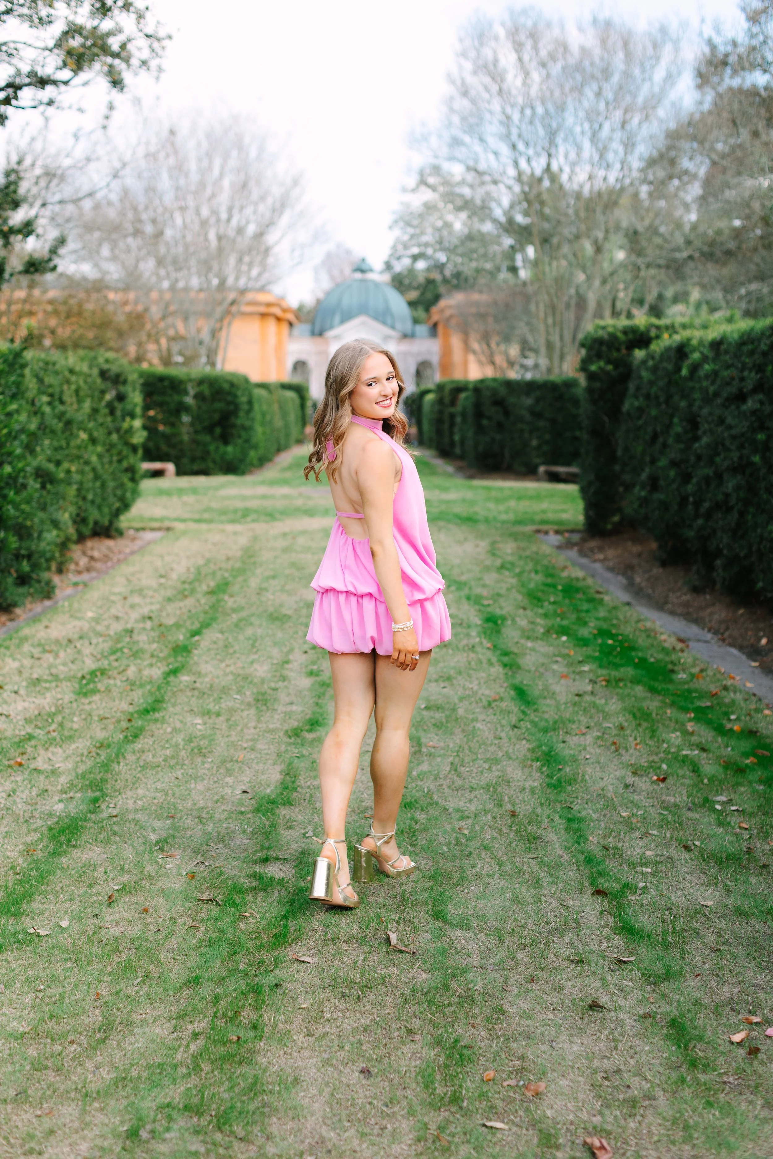 A woman wearing a pink dress and metallic high heels standing on a grassy pathway in a park with neatly trimmed bushes and trees, and a building with a dome in the background.