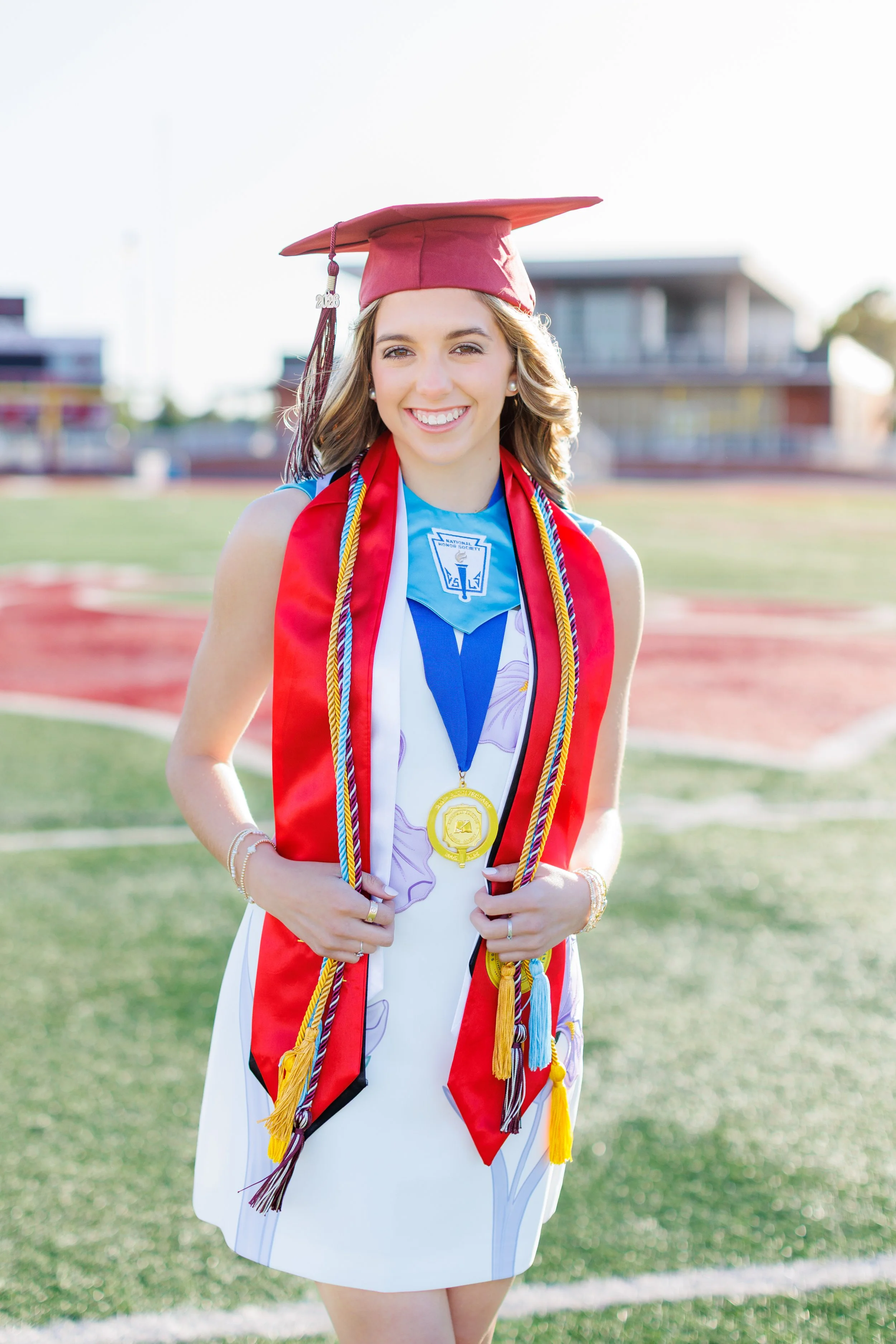 high school senior cap and gown session on field during golden hour. She wears honor cords and medals around her neck.