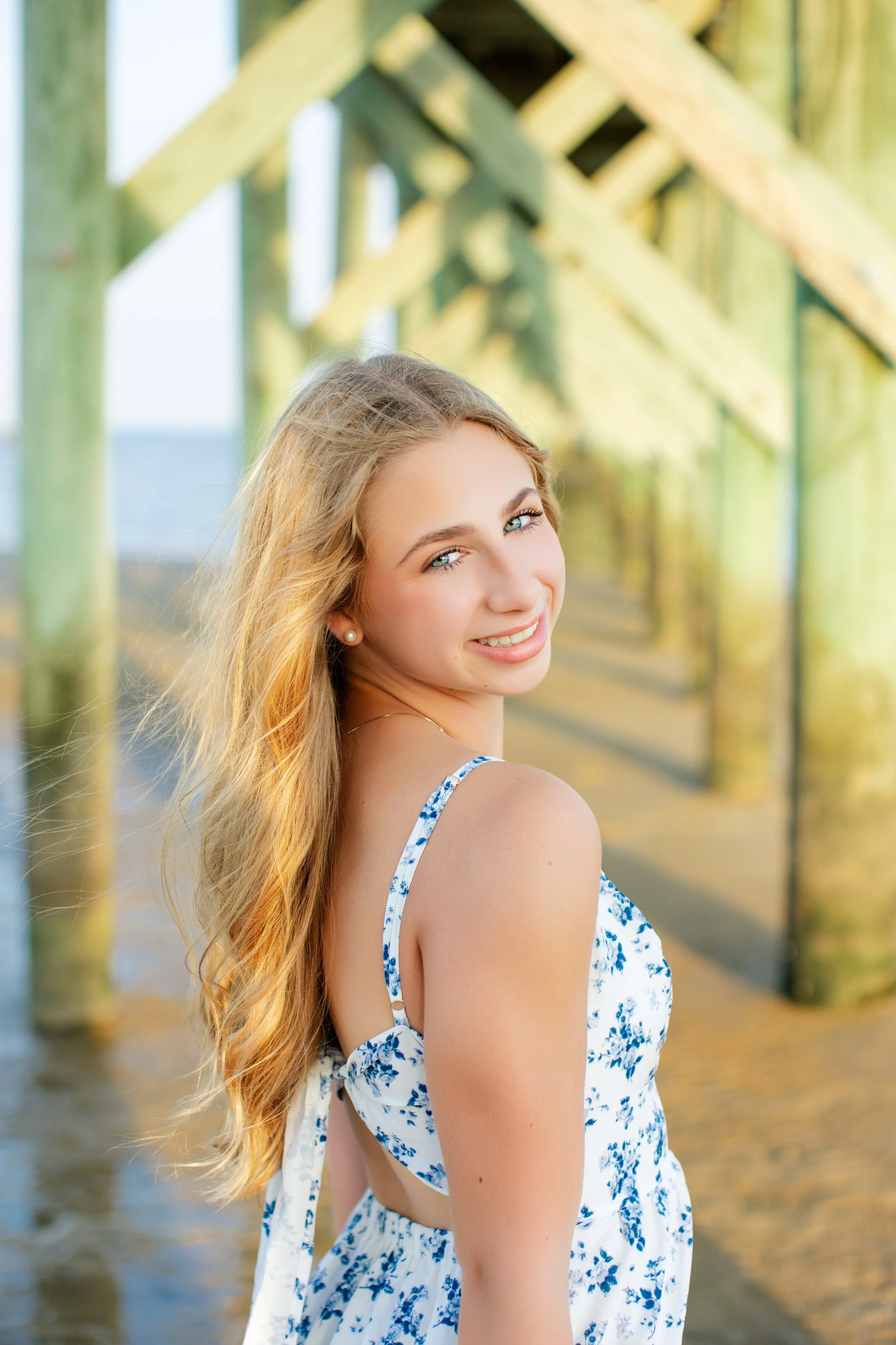 A young woman with long blonde hair and blue eyes, smiling, standing under a wooden walkway outdoors during daytime.