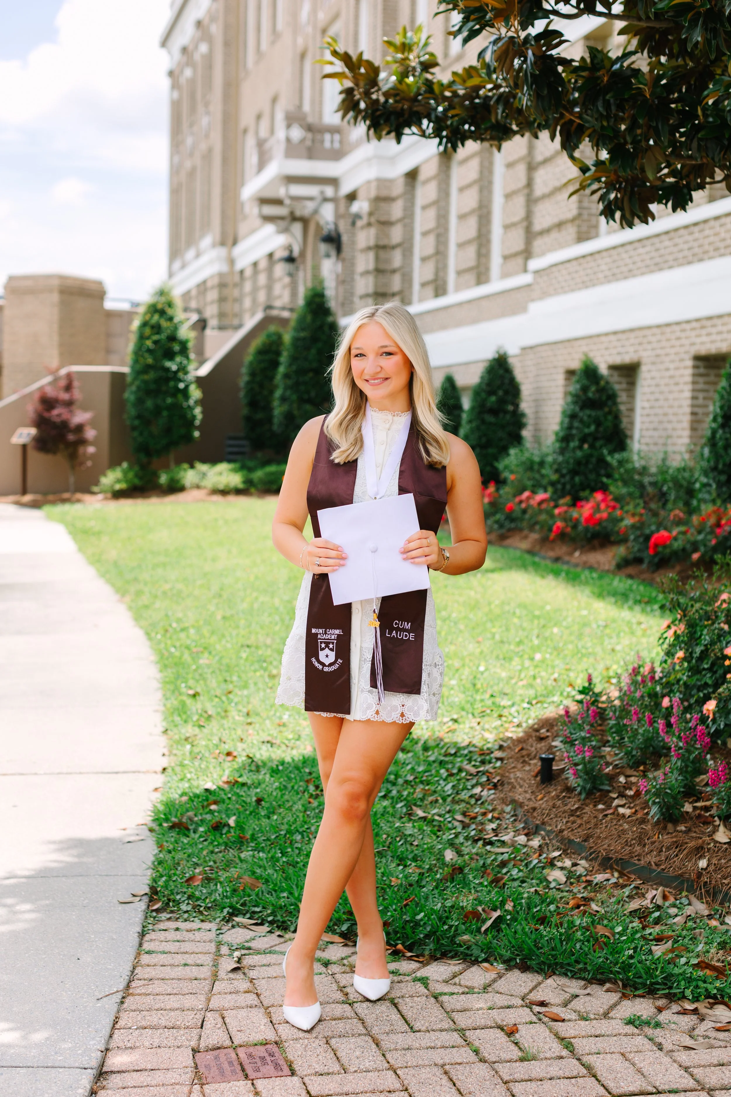 Young woman in a graduation cap and gown, holding a diploma, standing outside on a sidewalk with a landscaped garden and multi-story brick building in the background.