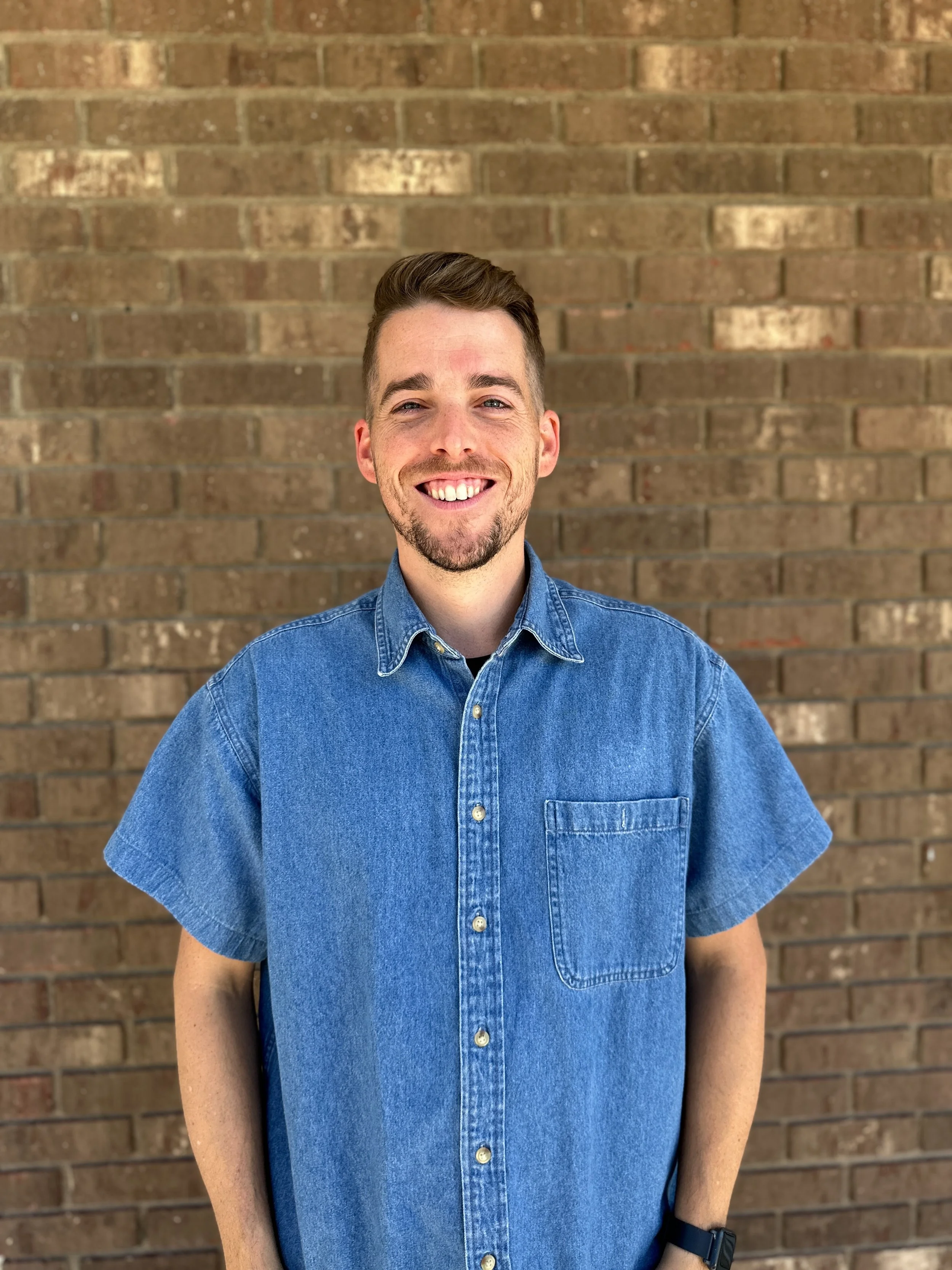 A young man with short brown hair and a beard smiling, wearing a denim short-sleeved shirt, standing in front of a brick wall.