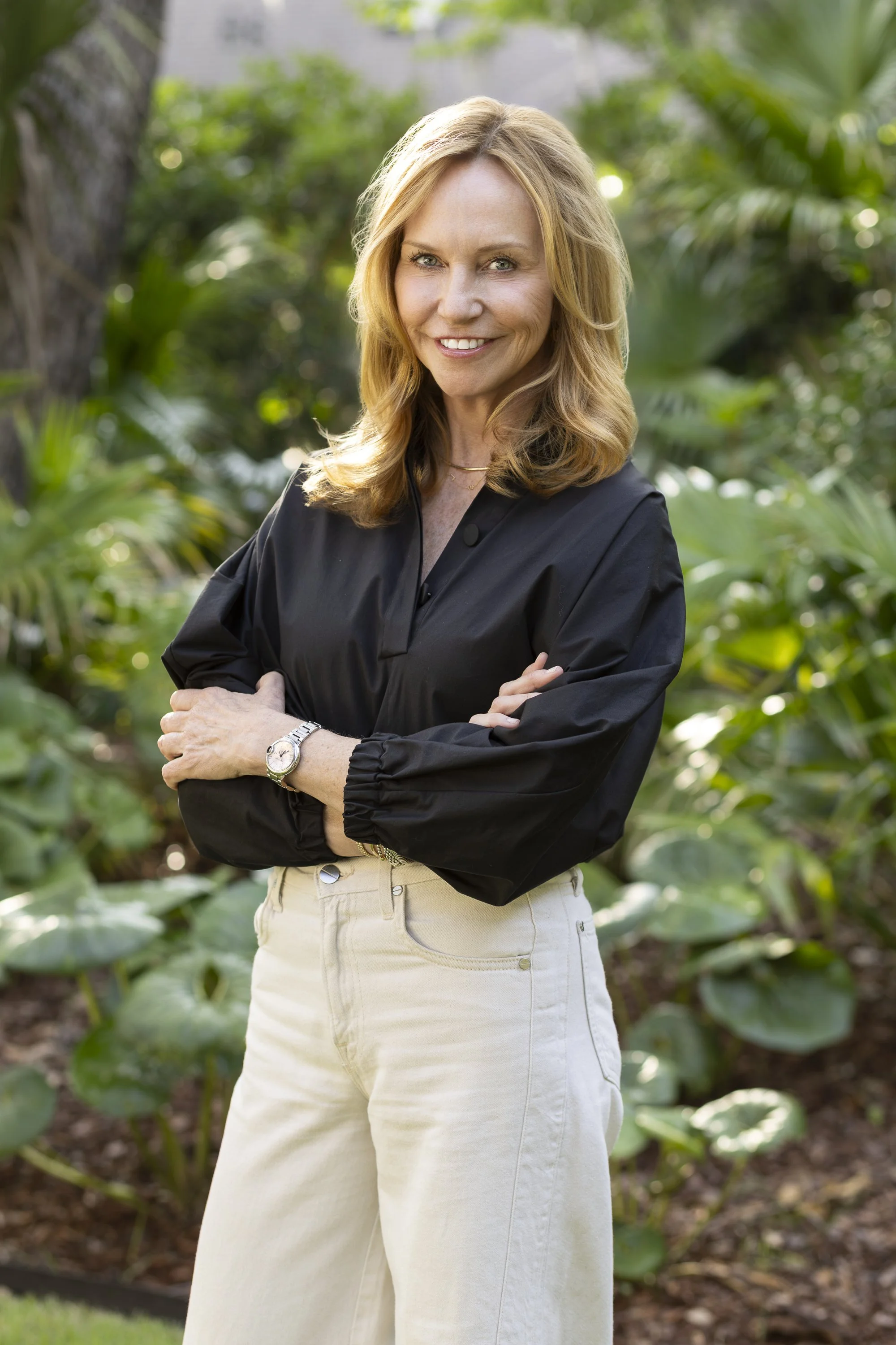 A woman with shoulder-length blonde hair smiling outdoors with green plants in the background, wearing a black long-sleeve blouse and cream-colored pants.