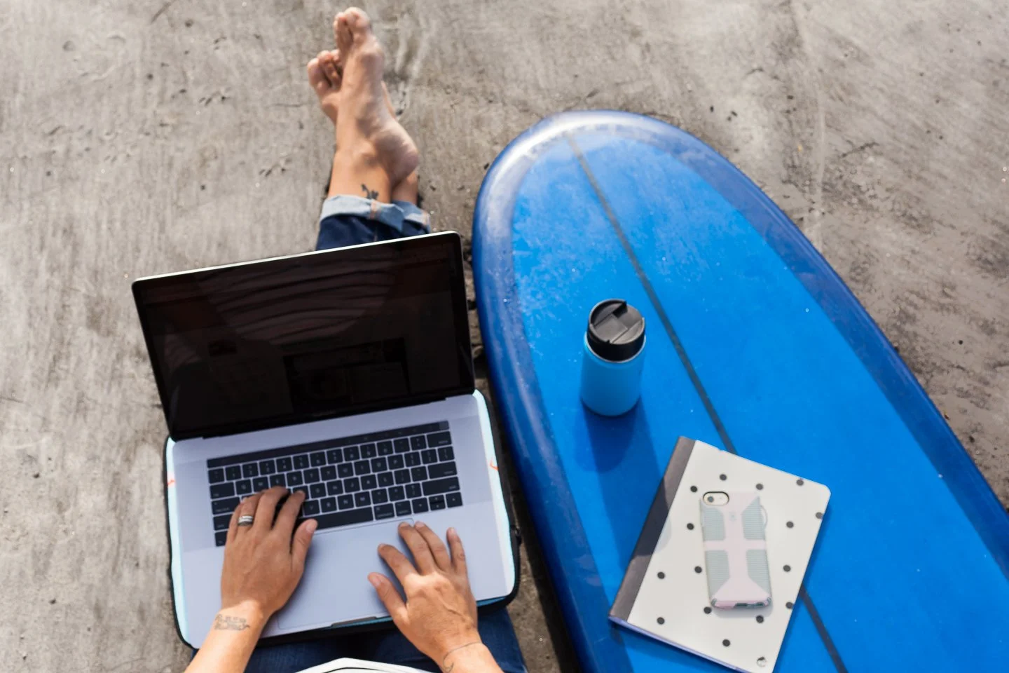 Person sitting on the ground working on a laptop, with a notebook and phone on a blue surfboard nearby, and a water bottle on the surfboard.