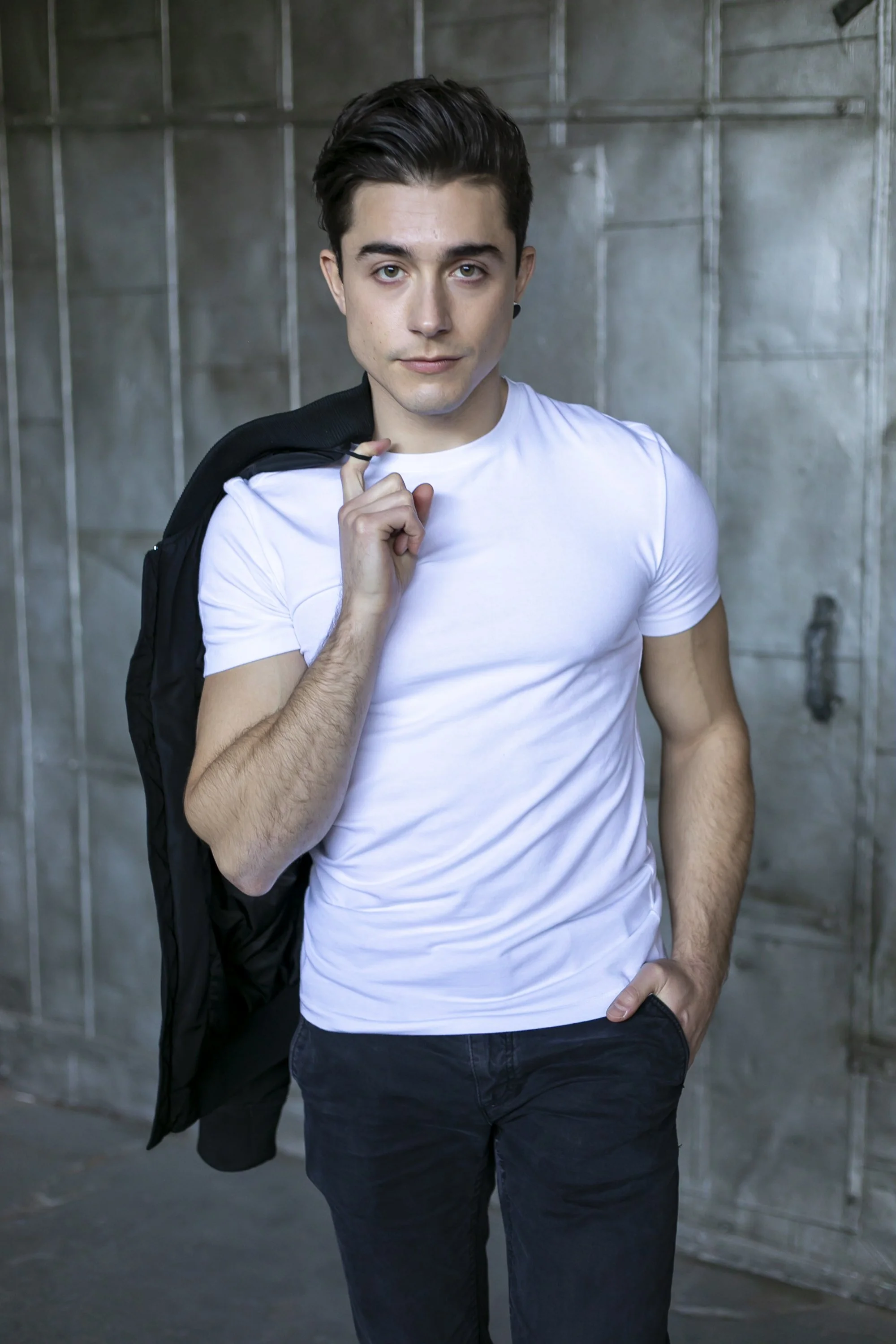 A young man with dark hair, wearing a white t-shirt and black pants, stands against a concrete wall. He is holding a black jacket over his shoulder with his right hand and has his left hand in his pocket. He gazes directly at the camera with a confid