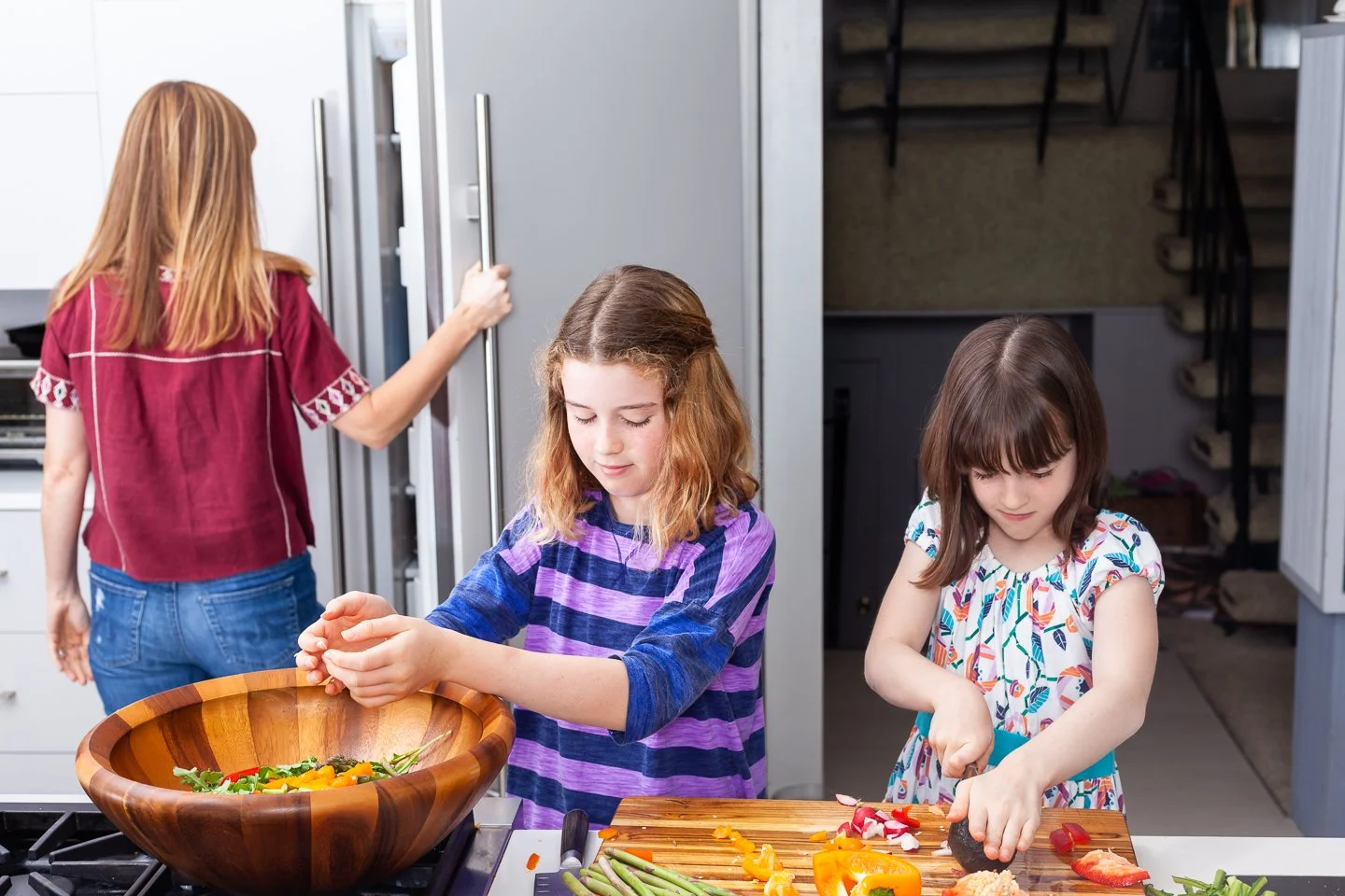 Two young girls chopping vegetables at a kitchen counter in a modern kitchen, with an adult woman opening a refrigerator in the background.